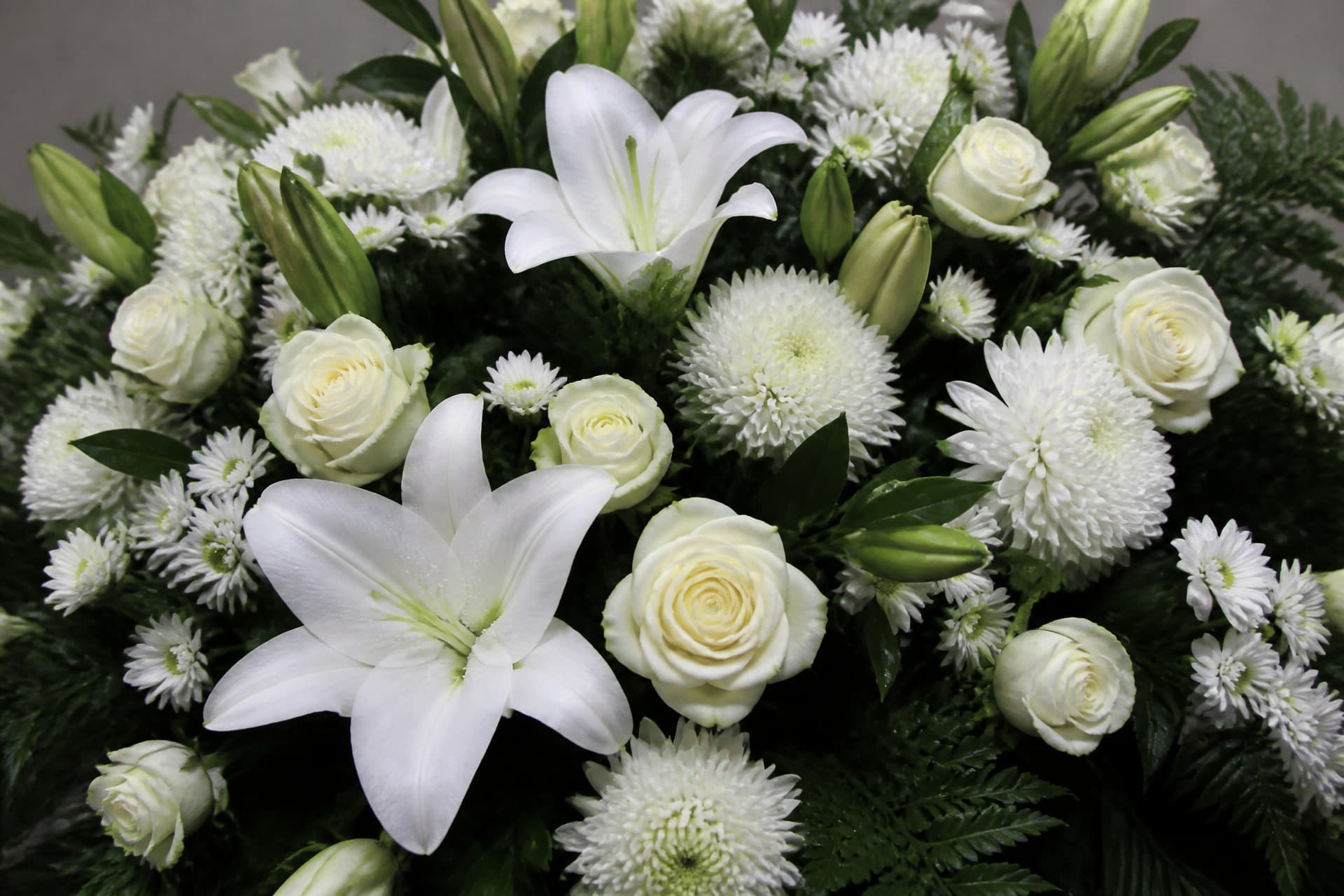 A close view of a white funeral arrangement filling the frame โ lilies, white roses, chrysanthemums and daisies against dark green foliage