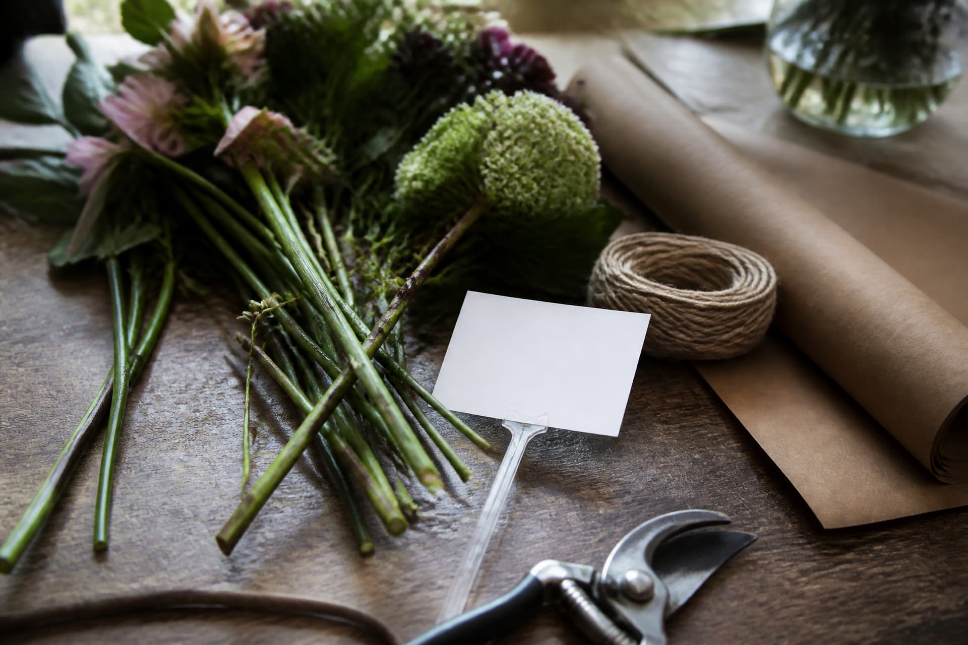 A florist's workbench with cut stems laid out, a roll of brown paper, twine and secateurs, and a blank white card clipped to a plastic stem pick