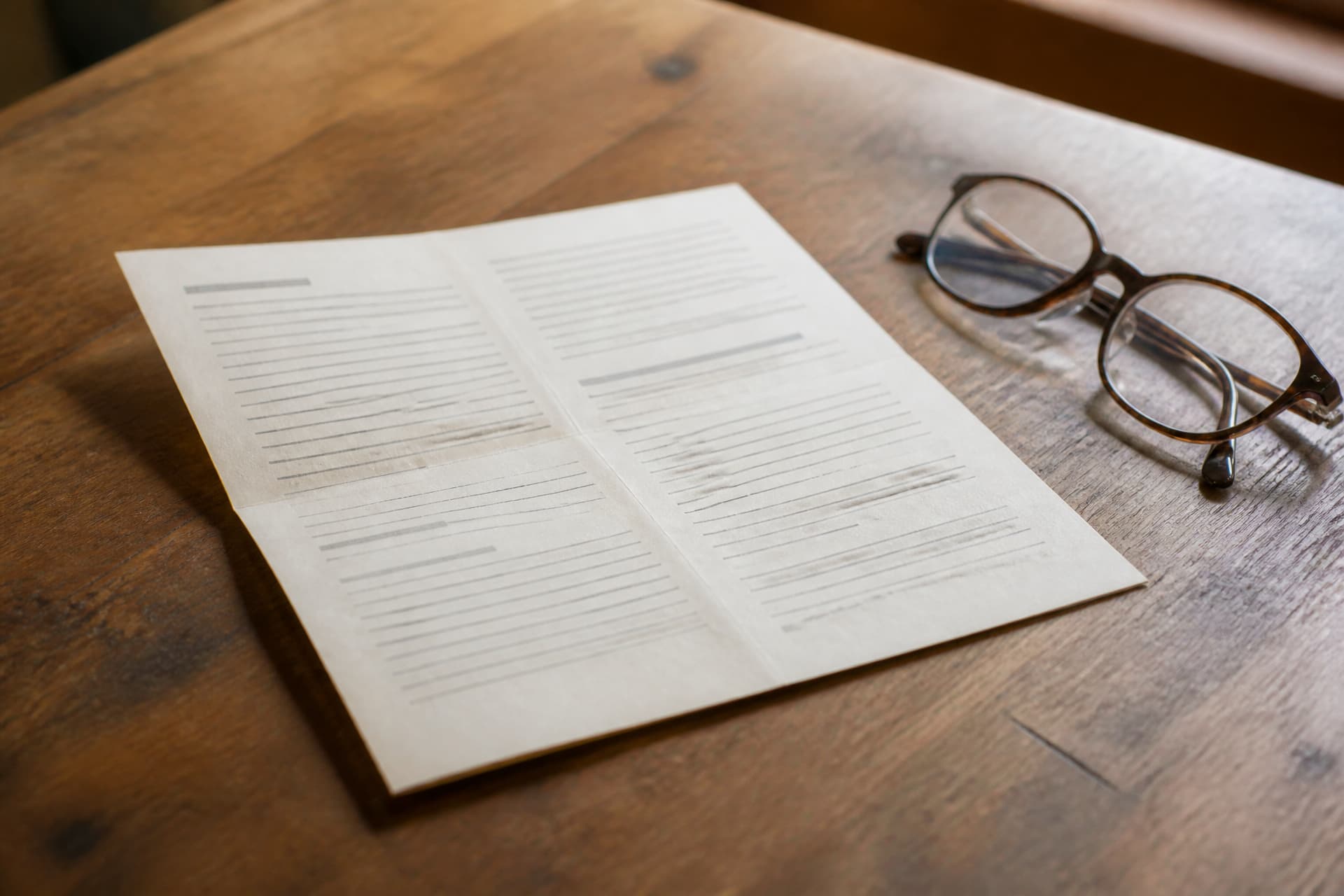 A folded printed leaflet open on a wooden table beside a pair of tortoiseshell reading glasses, its columns of type too soft to read