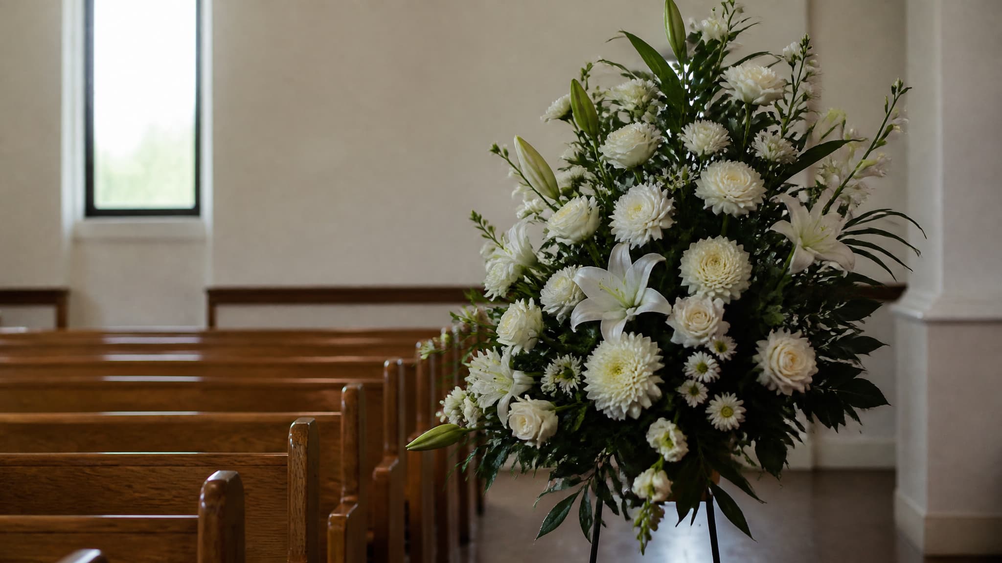 A large standing spray of white lilies, roses and chrysanthemums on a wire easel beside empty wooden pews in a plain chapel, daylight from a narrow window