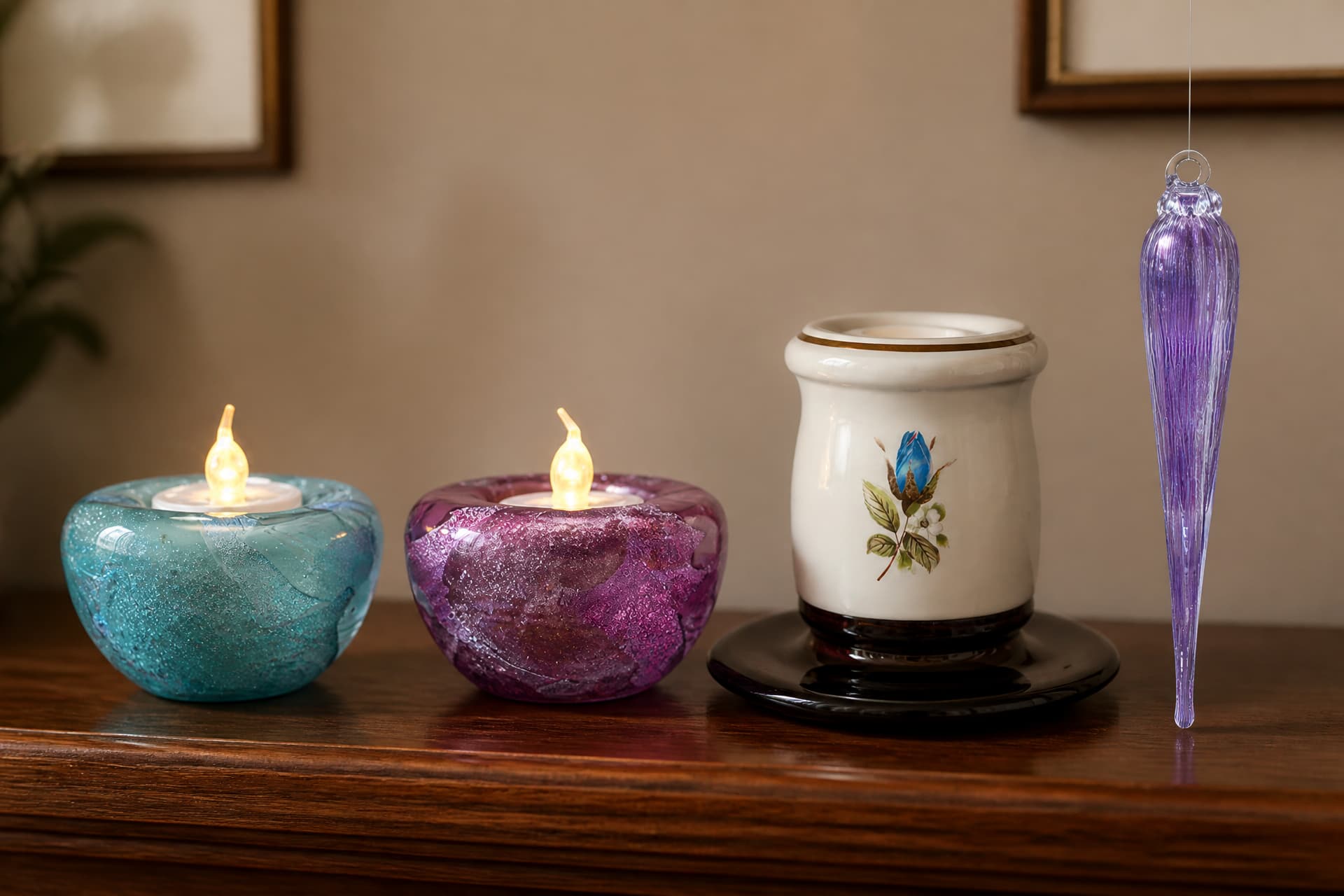 Two lit glass tealight holders in aqua and pink, a white candle holder painted with a blue rosebud on a dark saucer, and a slender violet glass icicle hanging beside them