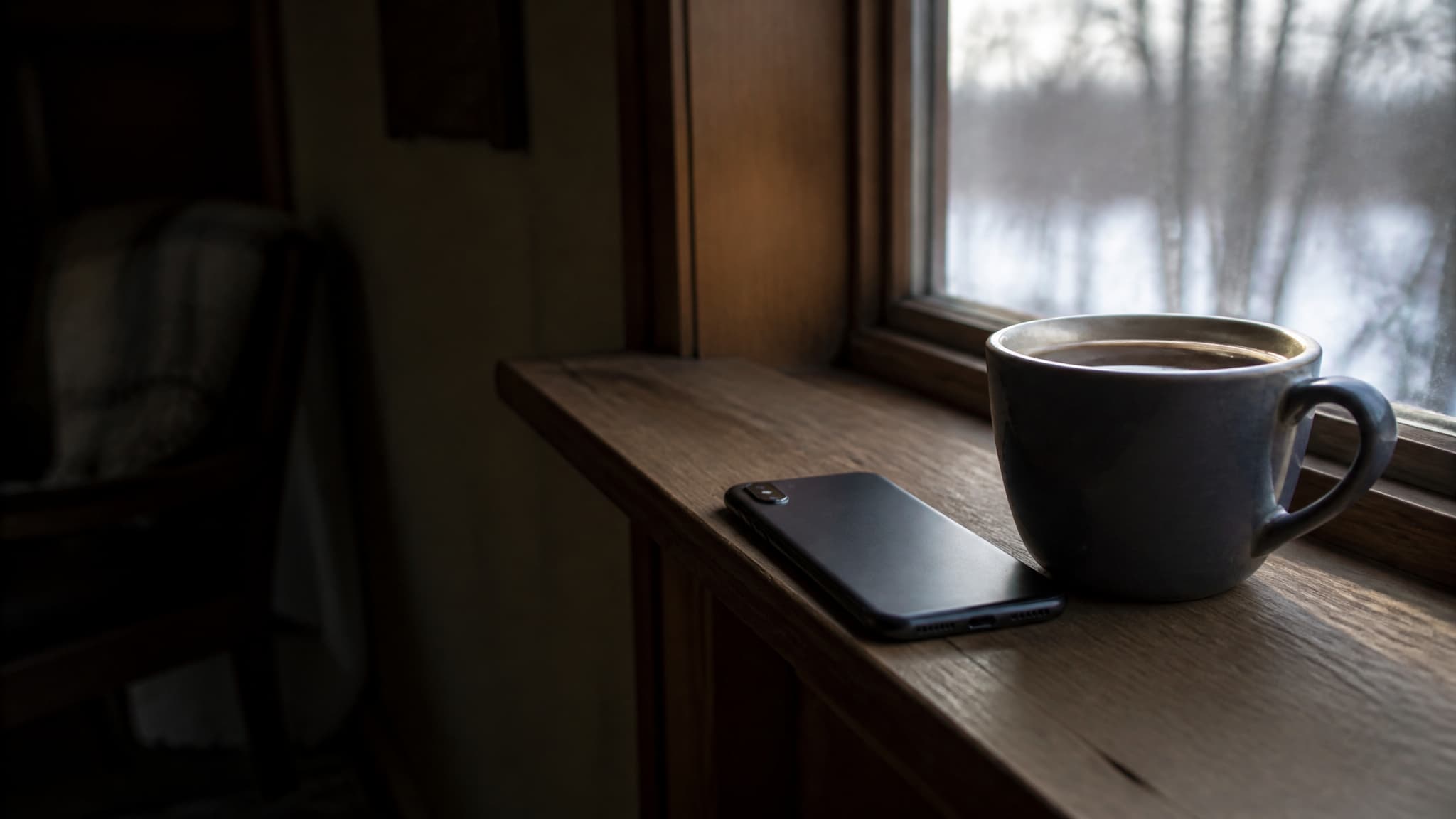 A phone face down on a windowsill beside a mug in low winter afternoon light, a quiet room behind