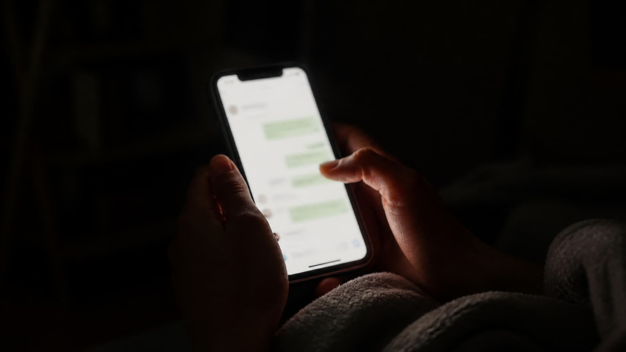 A phone held in two hands in an unlit room at night, the screen the only light in the frame and the thread on it too dim to read
