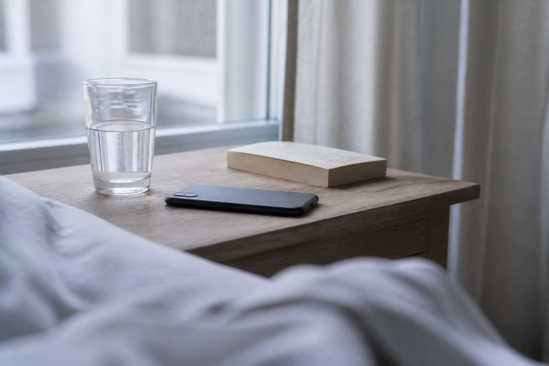 A phone lying face down on a wooden bedside table in pale early-morning light, beside a glass of water and a paperback, rumpled bed linen in the foreground