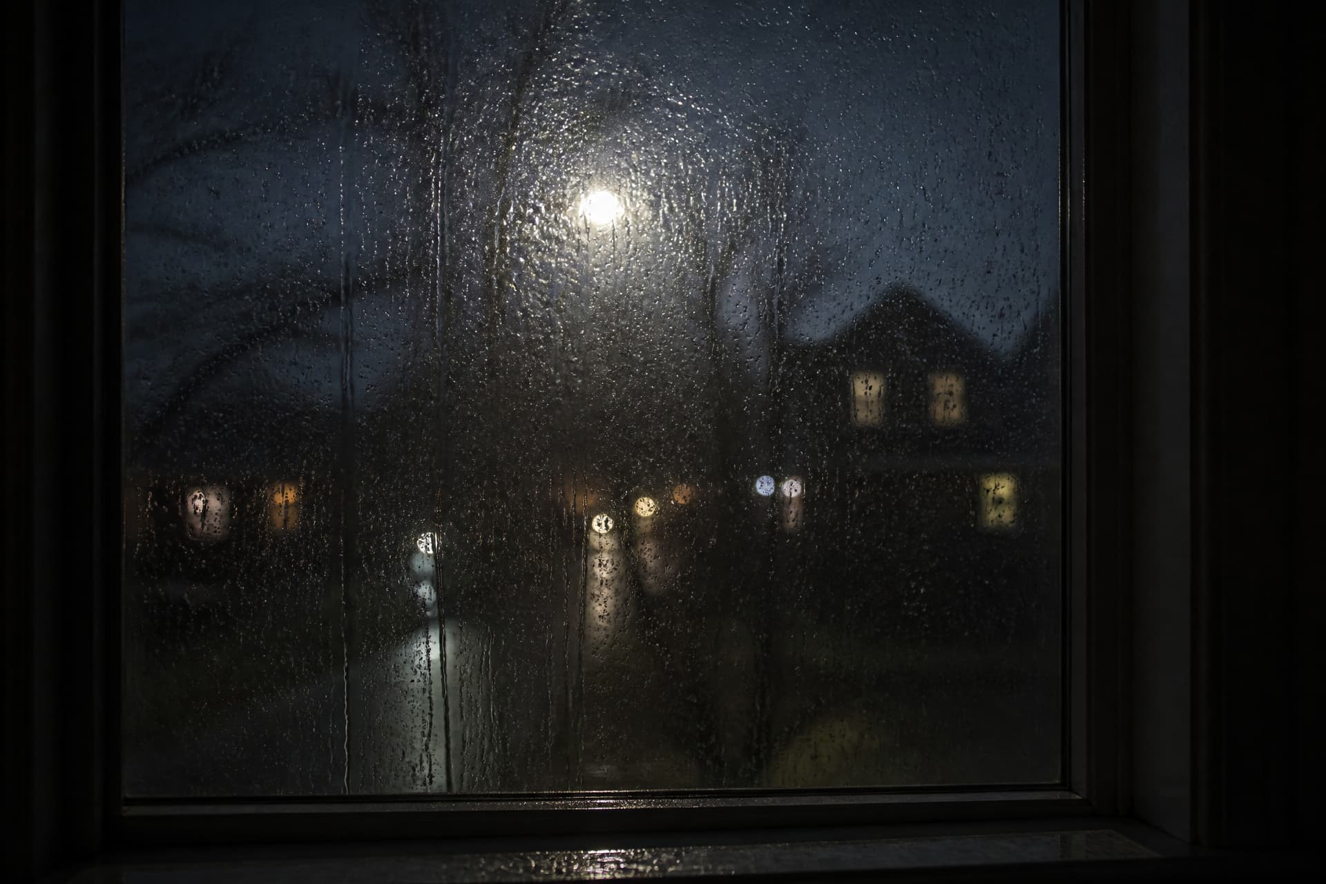 A rain-streaked window at night seen from inside a dark room, a streetlight and a few lit house windows blurred beyond the running water