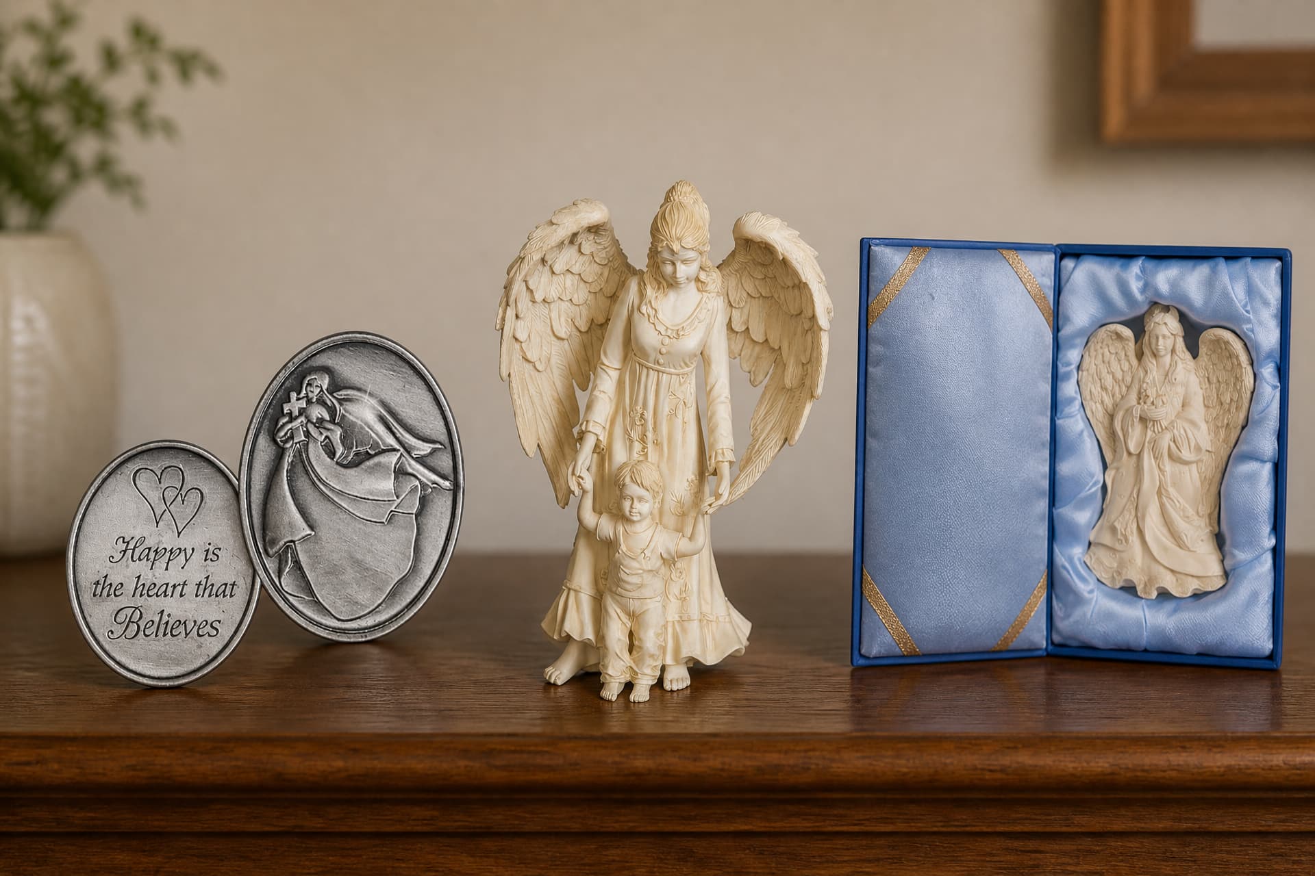 Two oval pewter comfort tokens propped on a wooden shelf beside a cast angel figure holding a child's hands, and a smaller angel seated in an open blue satin gift box