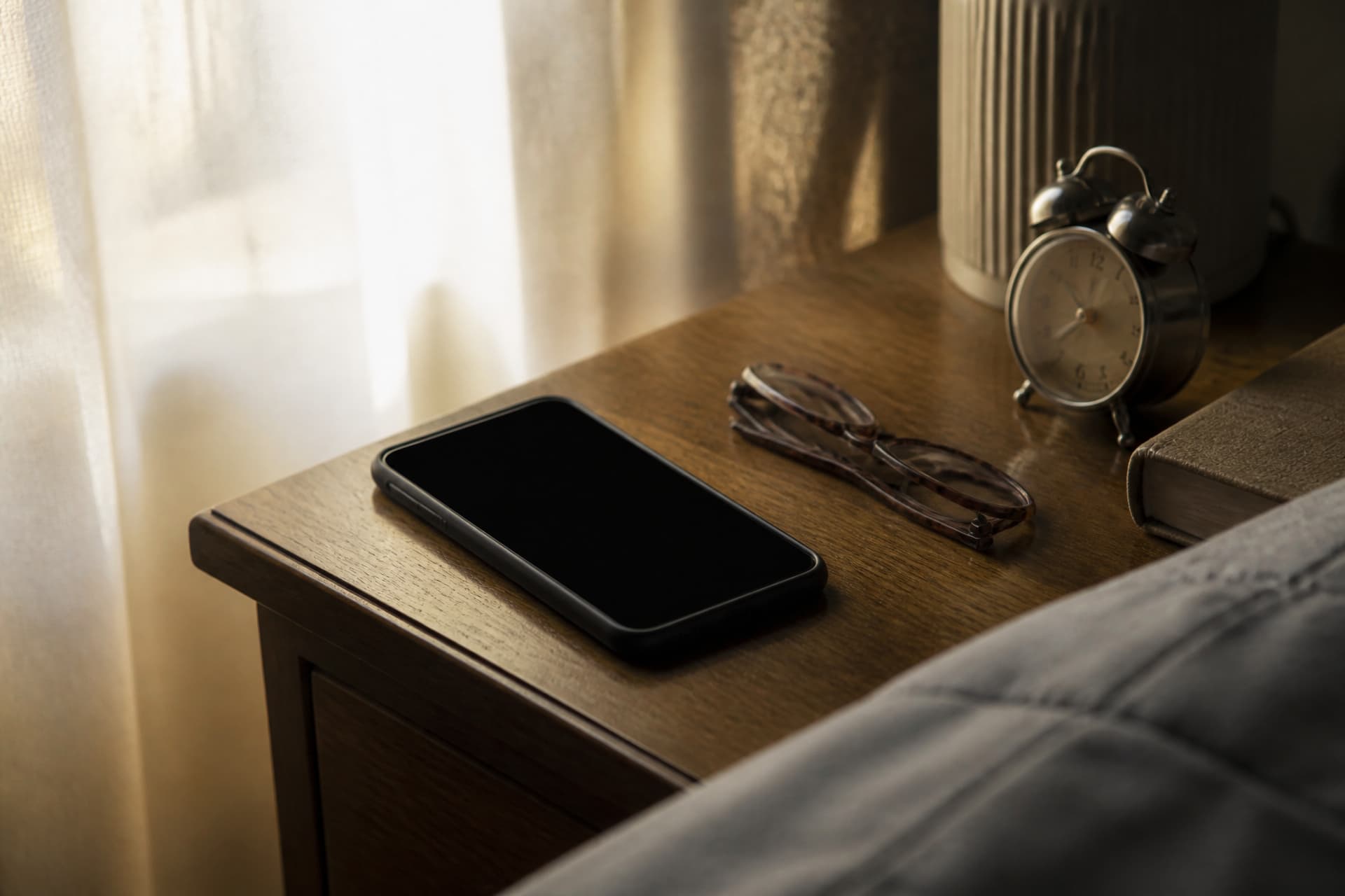 A phone face-up on a bedside table with a dark screen, an alarm clock and a folded pair of reading glasses beside it in early light