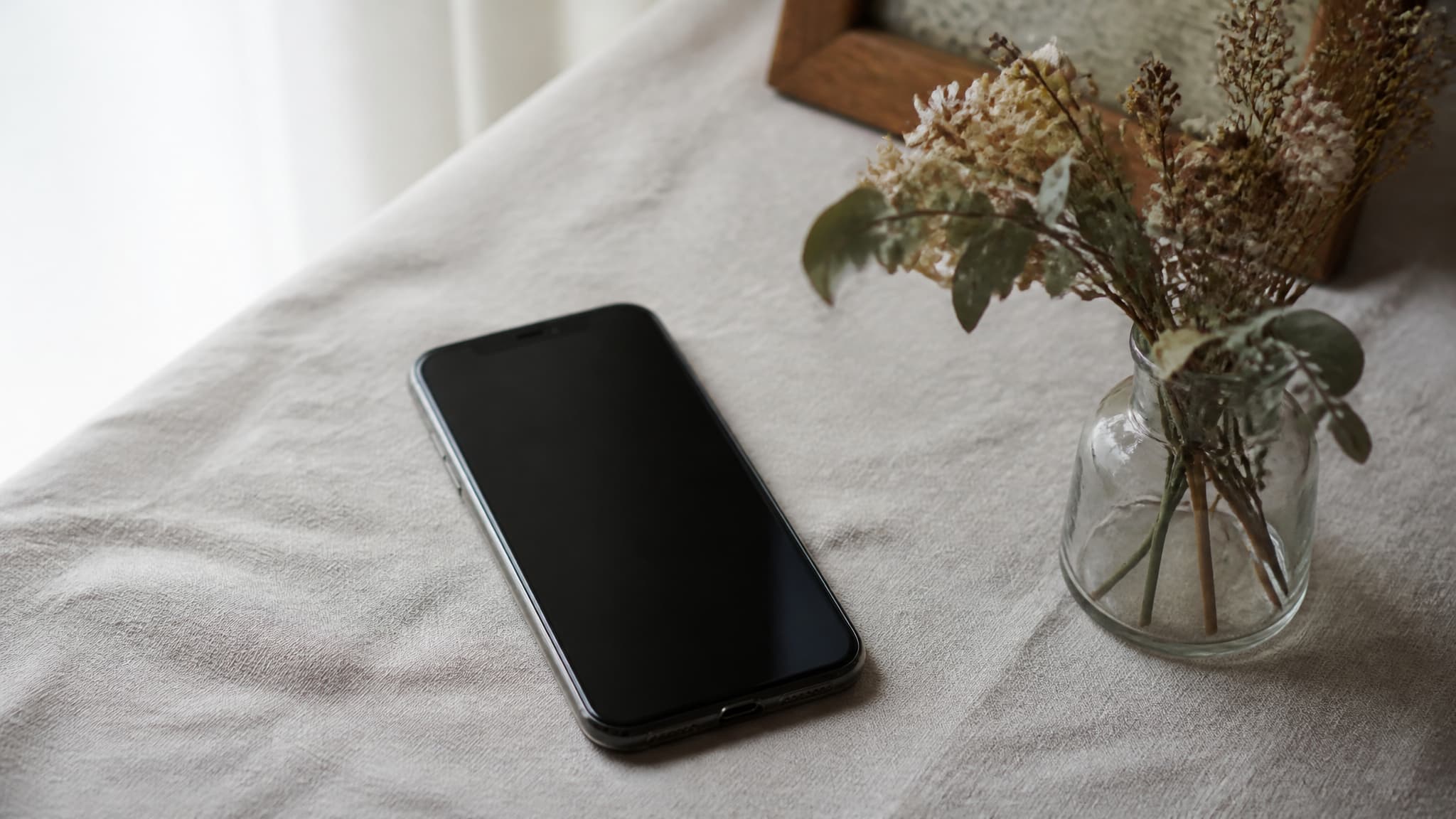 A phone lying face-up on a linen surface with a plain dark screen, beside a small vase of dried flowers in soft daylight