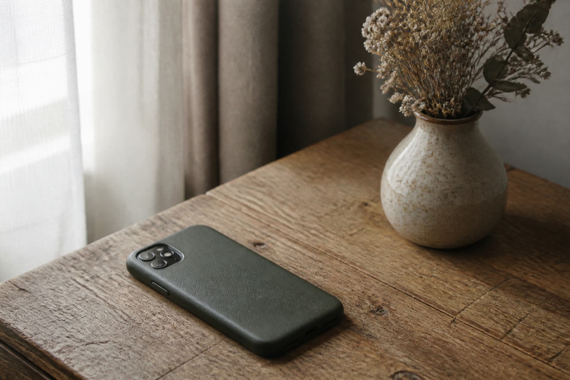A phone resting screen-down on a wooden table beside a small vase of dried flowers, soft window light from the left