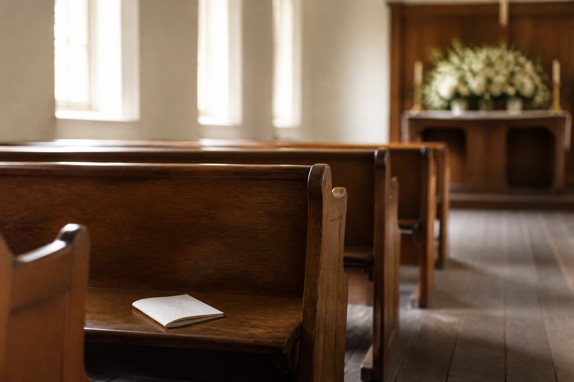 Rows of empty wooden chapel pews in soft daylight, with an order-of-service booklet left on one seat