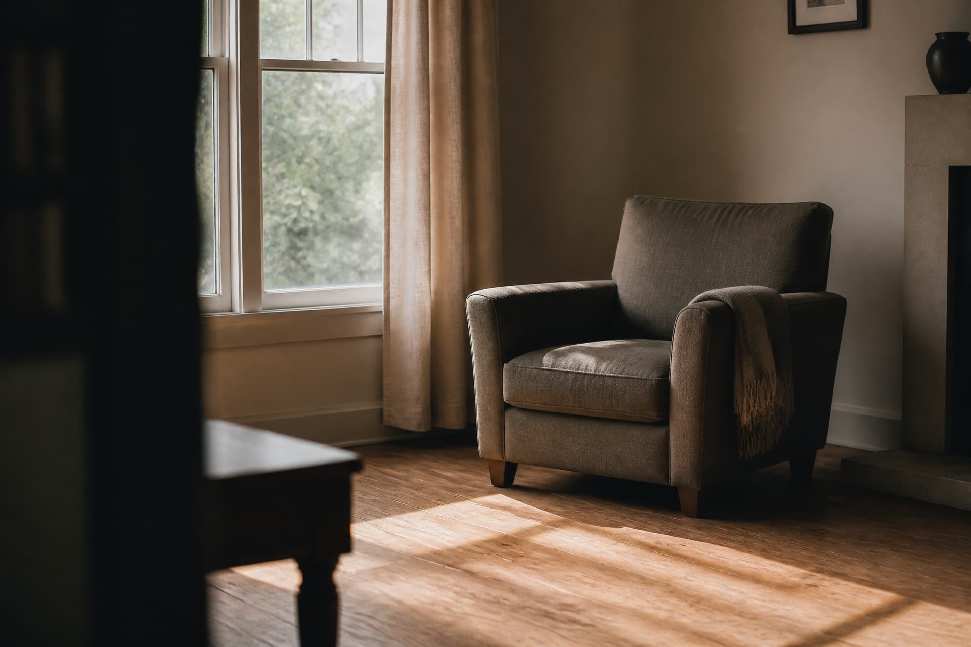 An empty grey armchair beside a bright window in a quiet living room, a fringed throw over one arm, afternoon light falling in bars across the wooden floor