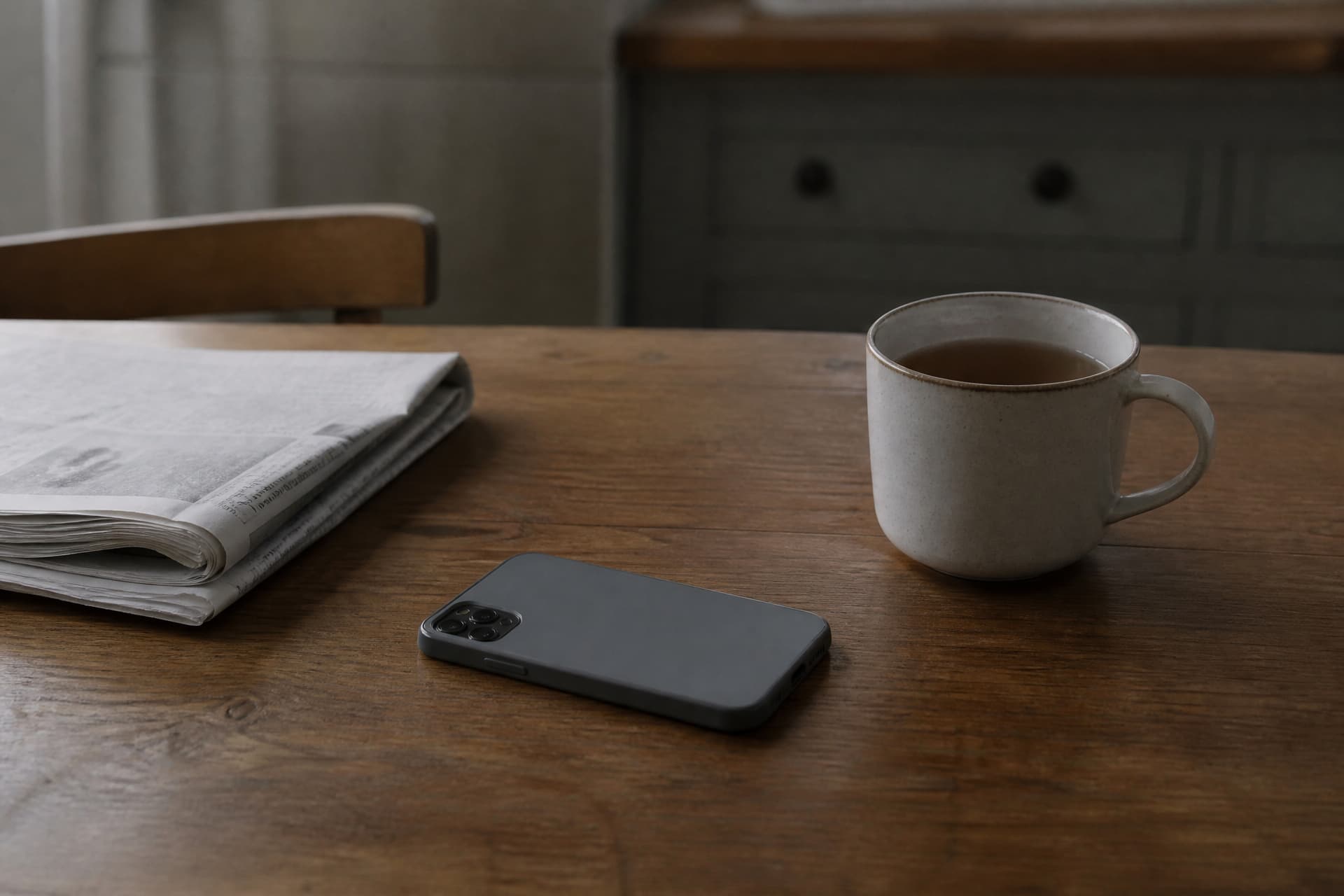 A phone lying face down on a wooden kitchen table beside an untouched mug of tea gone cold, a folded unopened newspaper at the edge of the frame