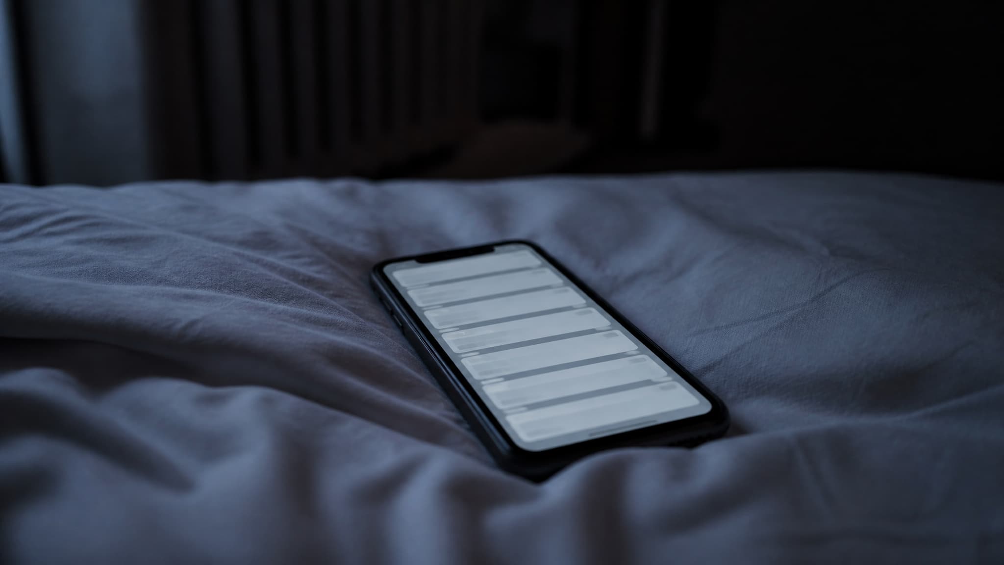 A phone lying face up on a rumpled pale bed sheet in a dark room, the screen glowing with a column of stacked notification cards too small to read