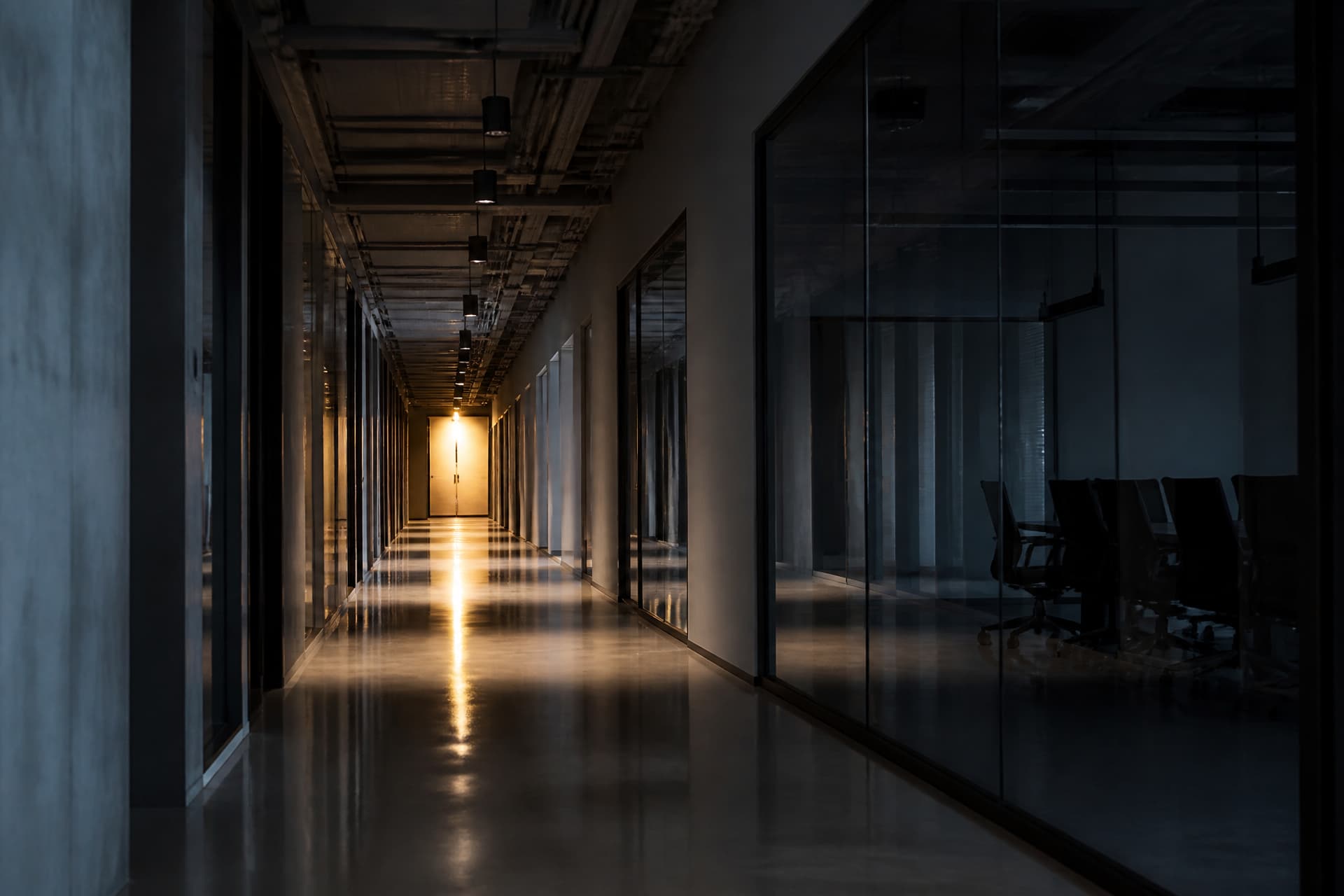 An empty office corridor before anyone has arrived, a dark glass meeting room along one side, a single warm light at the far end reflecting down the polished floor