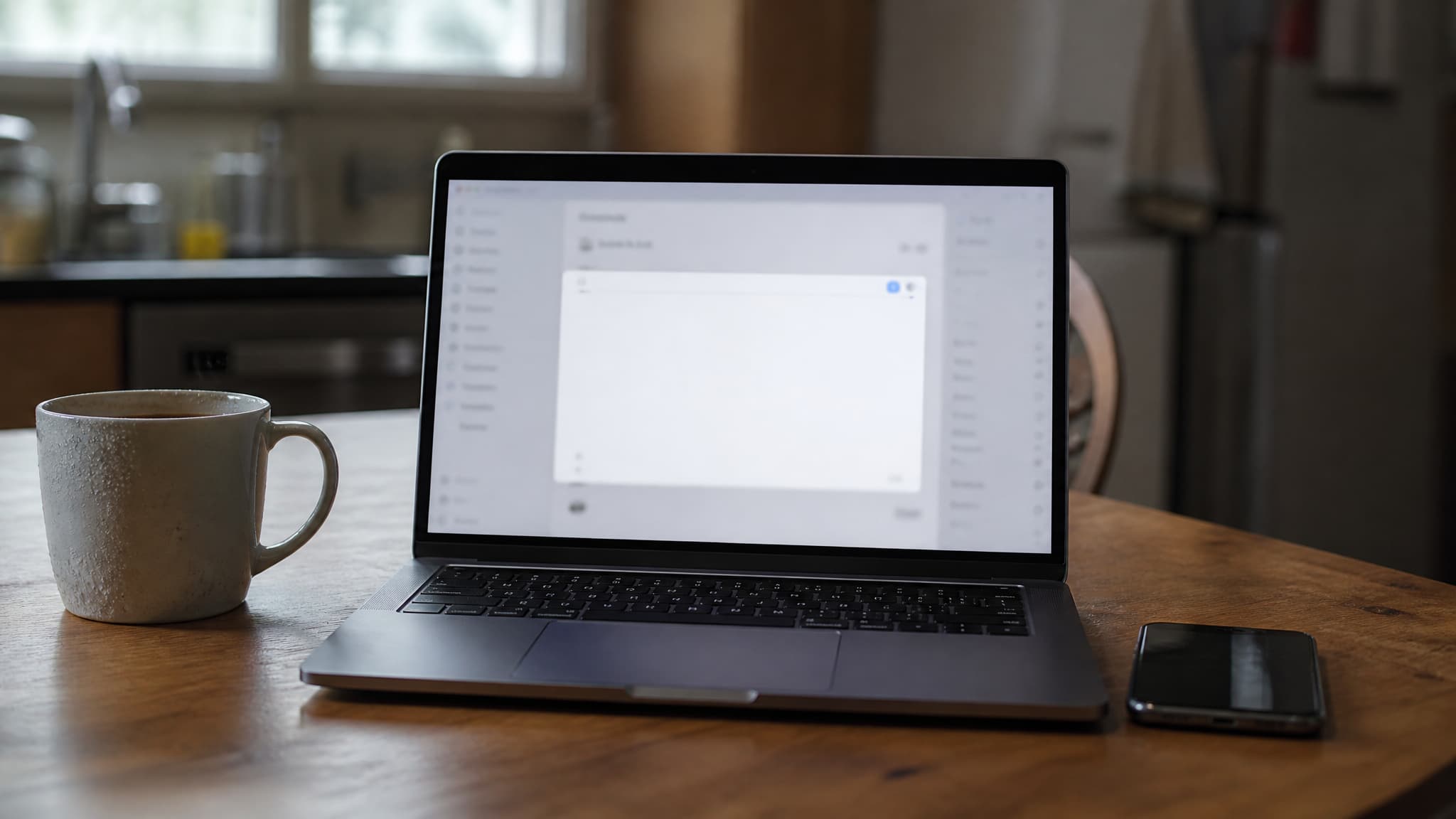 A laptop open on a kitchen table in early morning light showing a blank compose window, a mug of coffee and a phone beside it