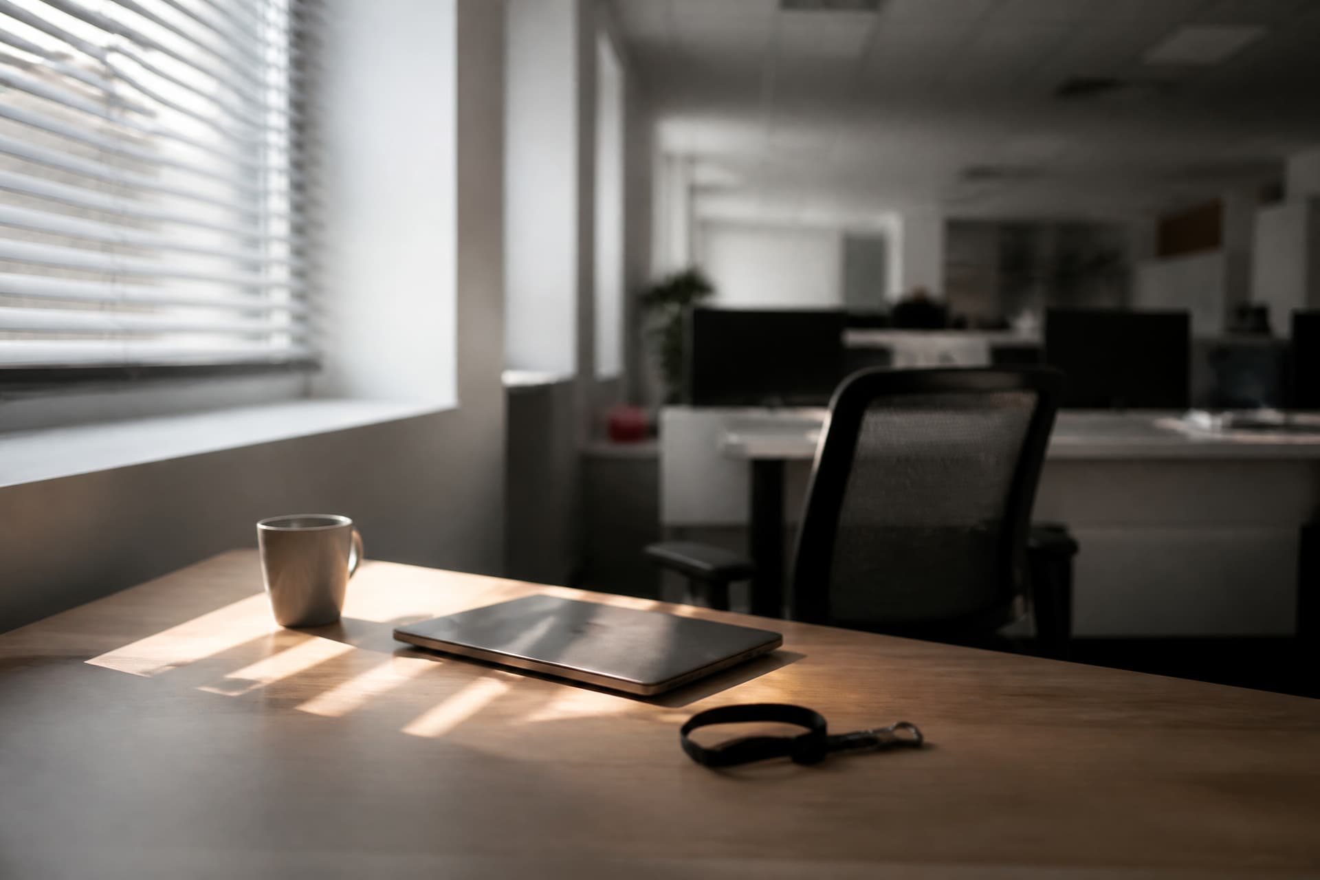 An office desk with an empty chair pushed back, a closed laptop, a lanyard coiled beside a mug, low morning light through blinds
