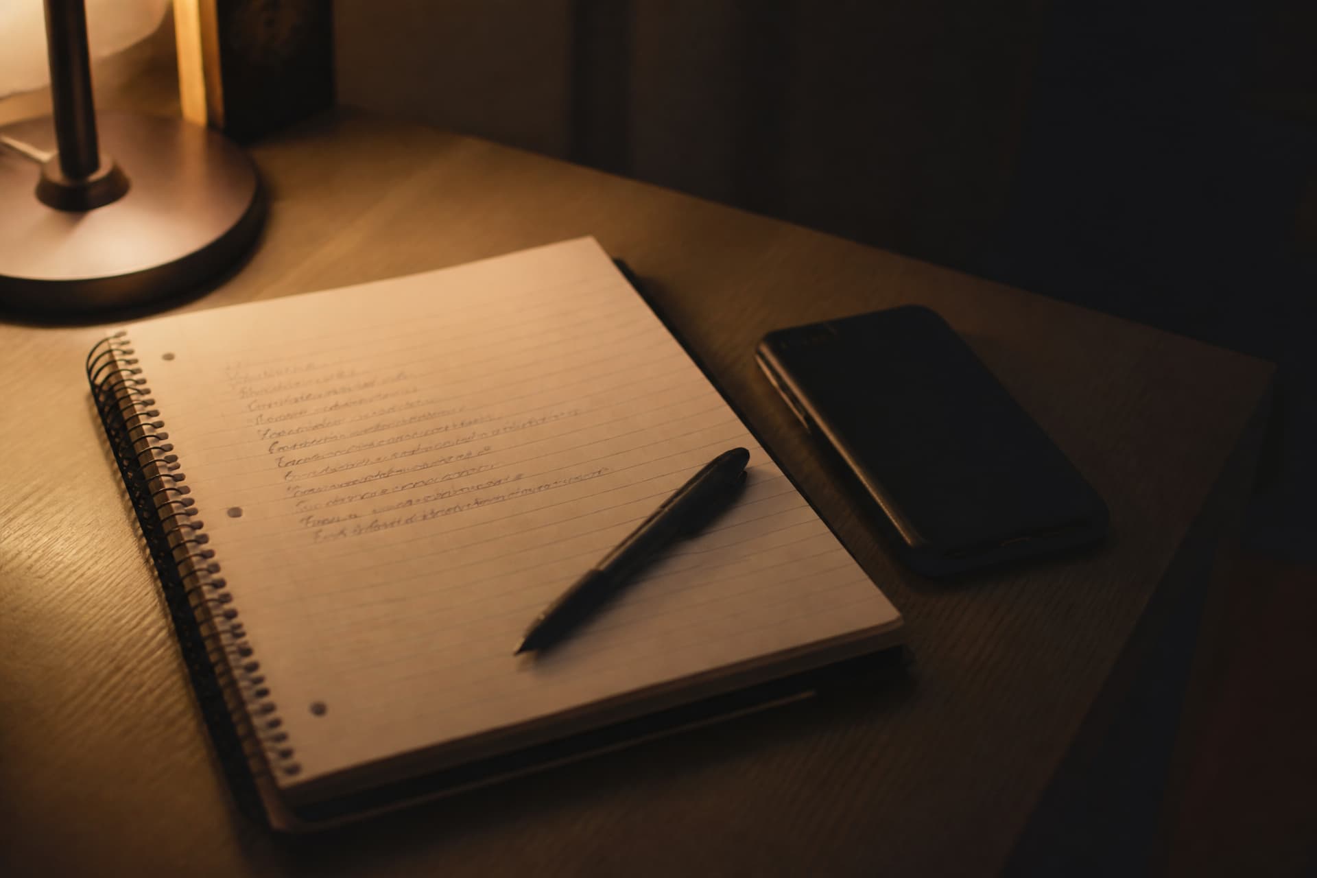 An open spiral notebook on a desk under a lamp, a short handwritten list on the page, a pen laid across it and a phone face down beside it