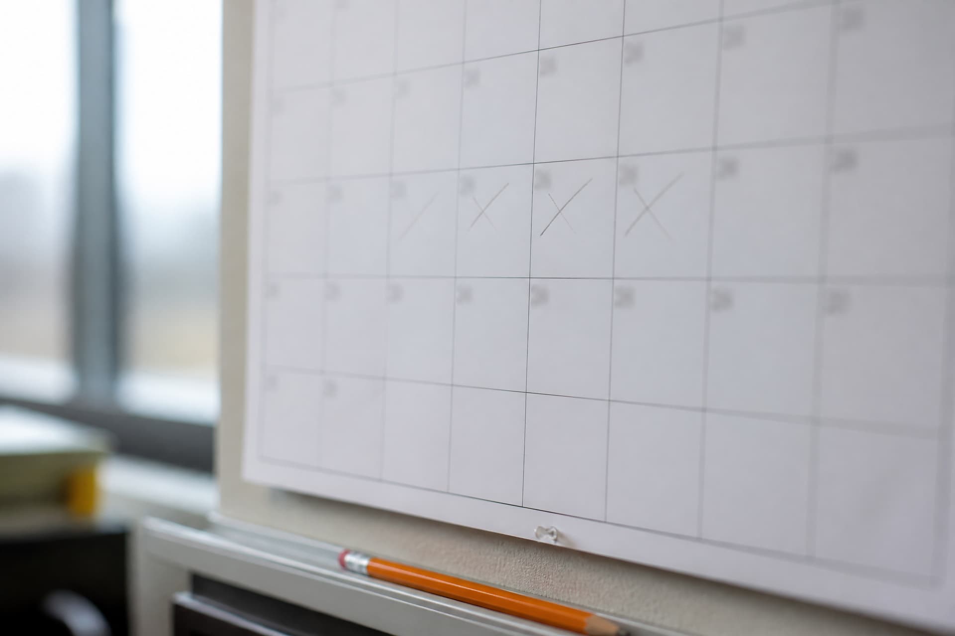 A paper wall planner pinned by an office window, its month grid blank except for four consecutive days crossed off in soft pencil