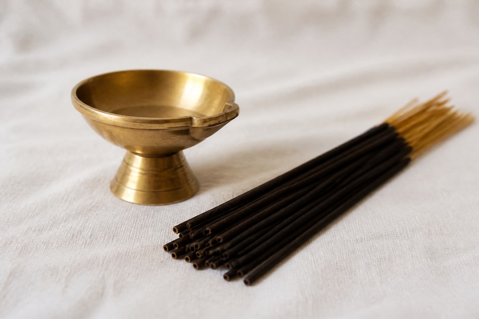A small plain brass oil lamp beside a loose bundle of unlit incense sticks resting on pale cloth