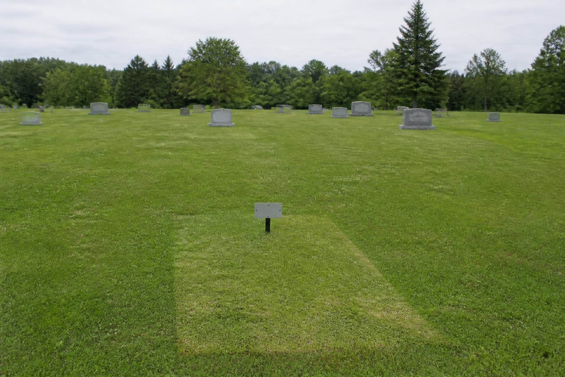 A newly settled grave plot in a lawn cemetery, still unmarked, with a small blank temporary marker stake at one end