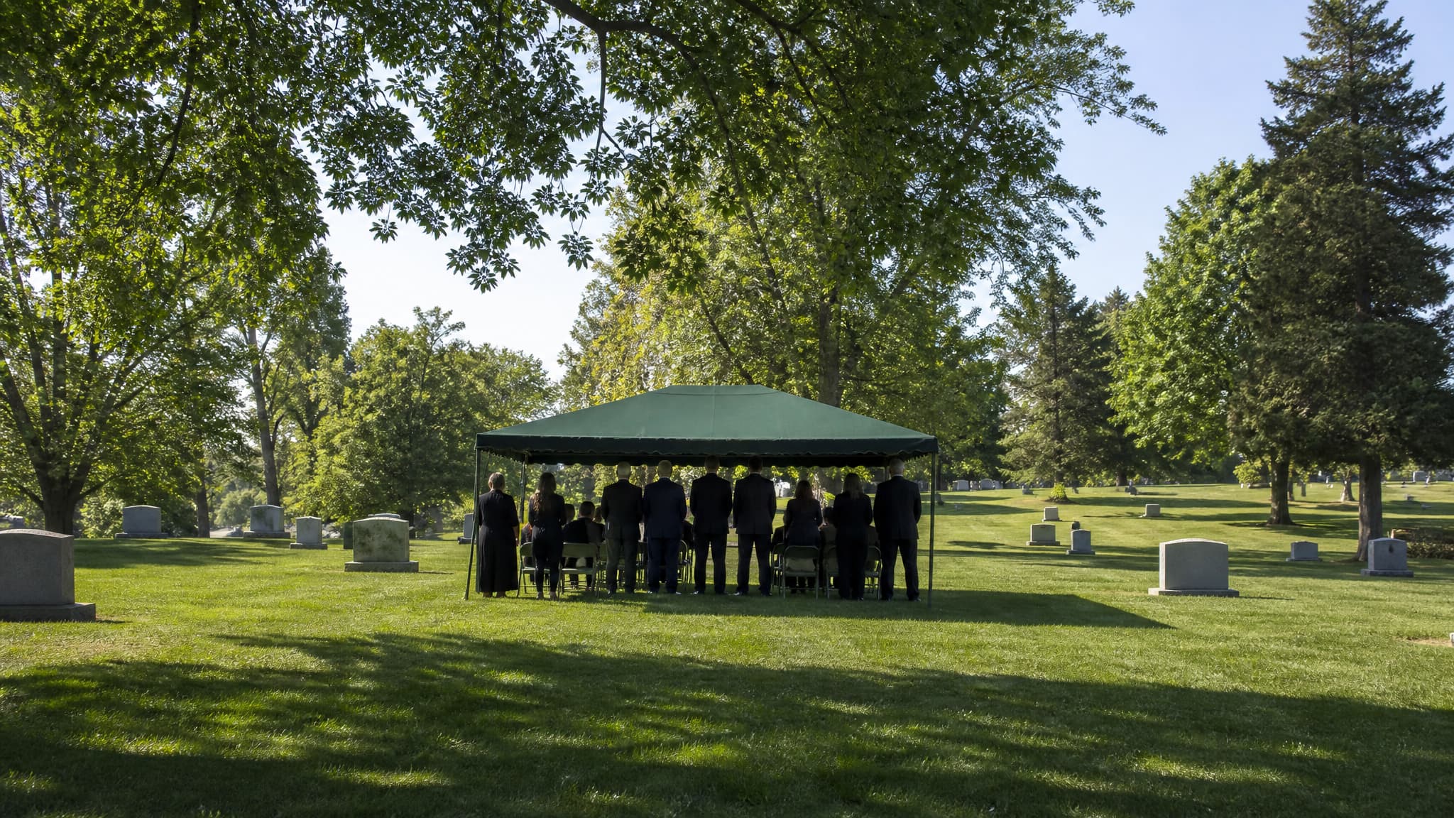 A small graveside service gathered beneath a green canopy on a cemetery lawn, seen from a distance behind the group