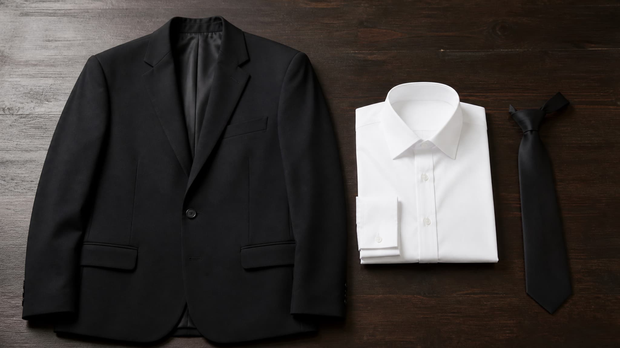 A plain black suit jacket beside a neatly folded white shirt on a dark wooden surface