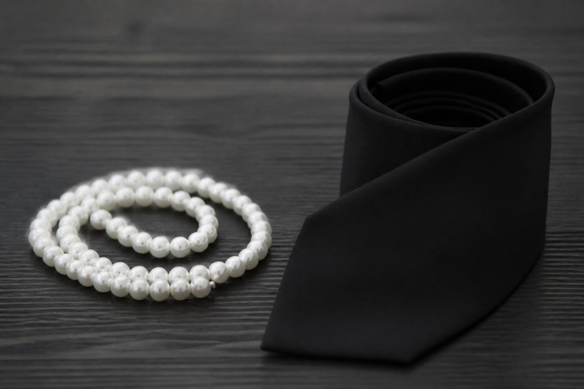 A single strand of white pearls coiled on dark wood beside a plain matte black necktie rolled into a coil