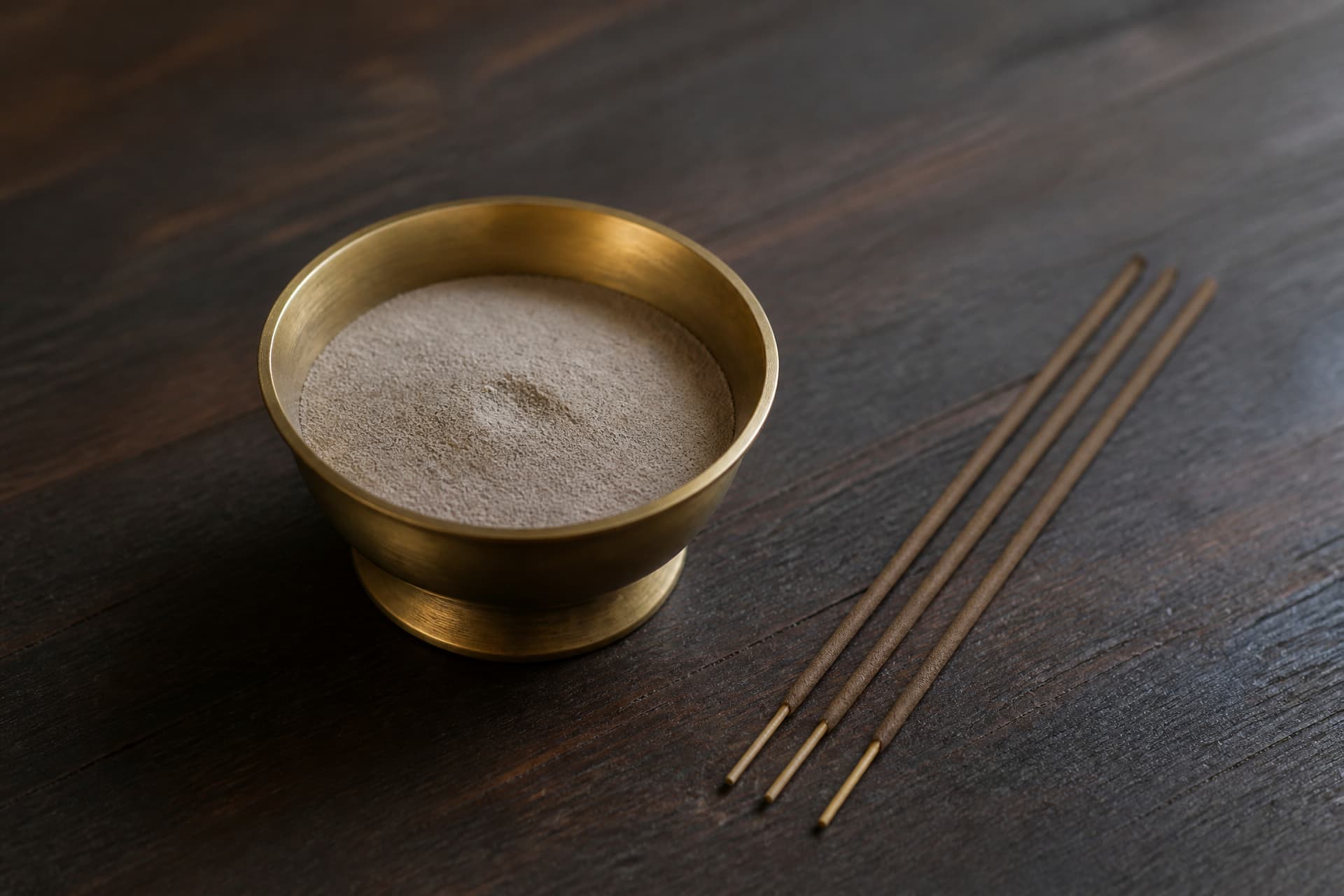 A small brass incense burner holding fine grey ash beside a few unlit incense sticks on a dark surface