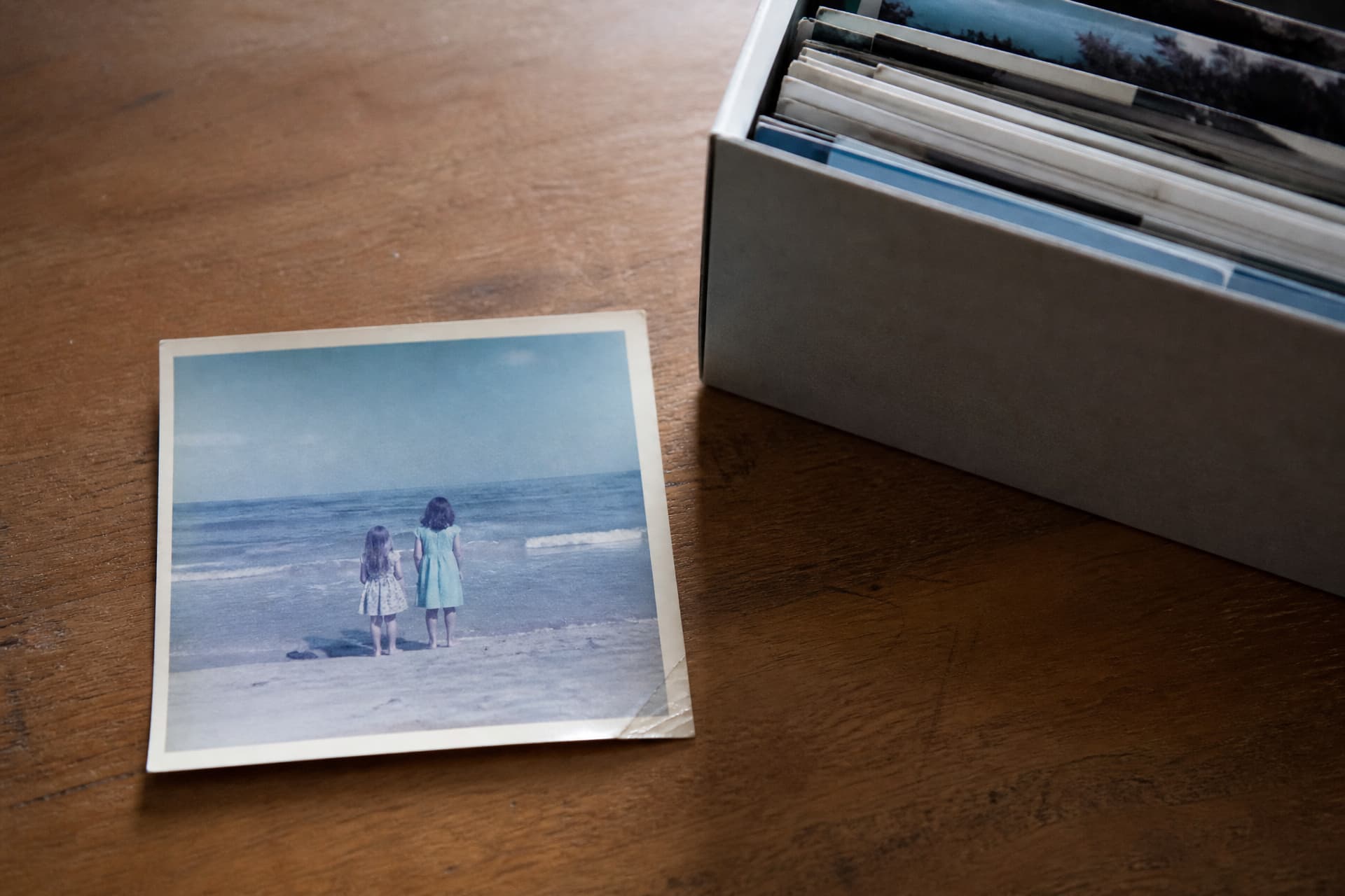A faded color snapshot lying face up on a wooden table beside an open box of loose prints, showing two girls in summer dresses standing at the water's edge with their backs to the camera, one corner of the print creased