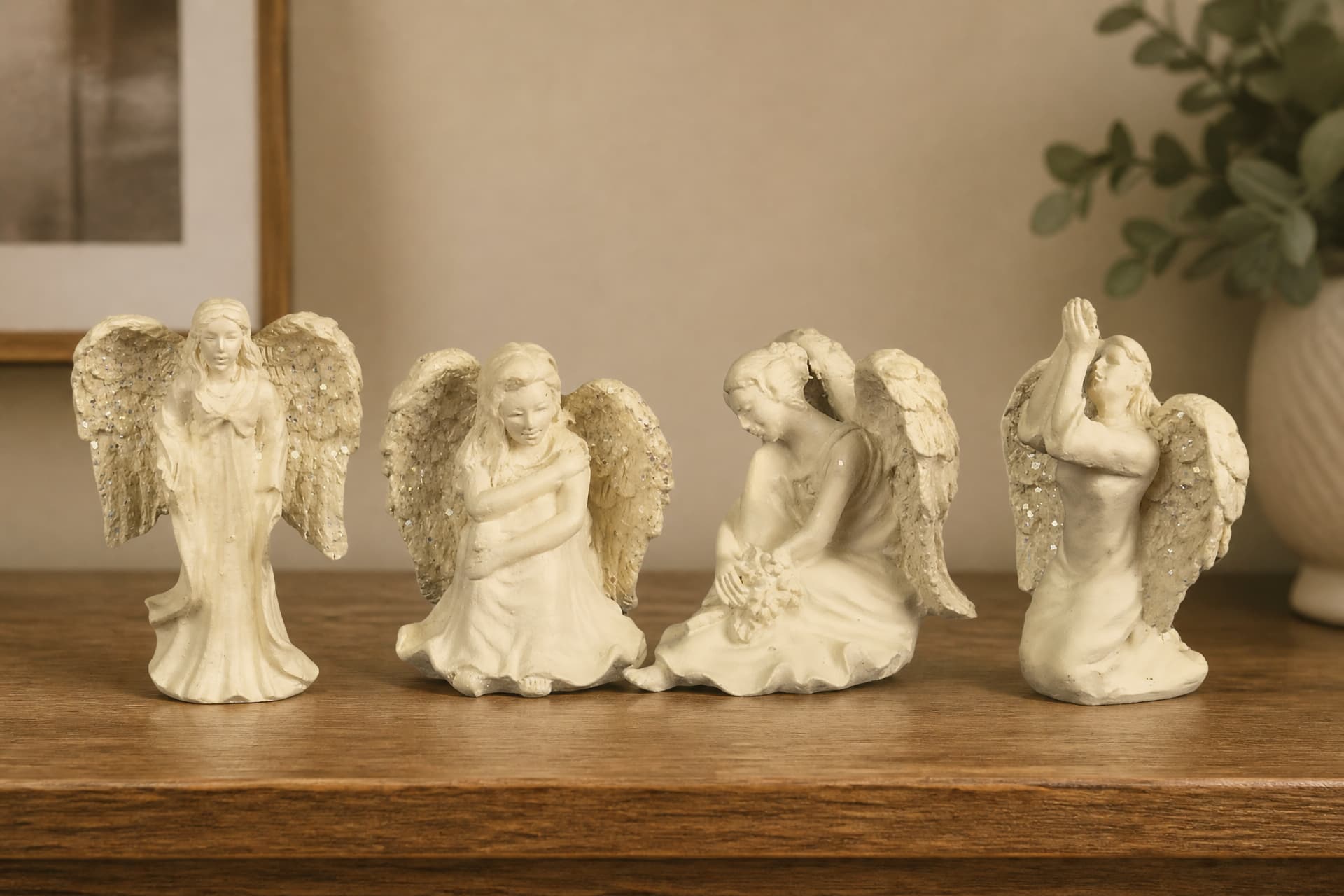 Four small cream cast-stone angel figures in a row on a wooden shelf, one standing with hands clasped, one kneeling with arms crossed, one seated with her head bowed over a posy, one kneeling with both arms raised