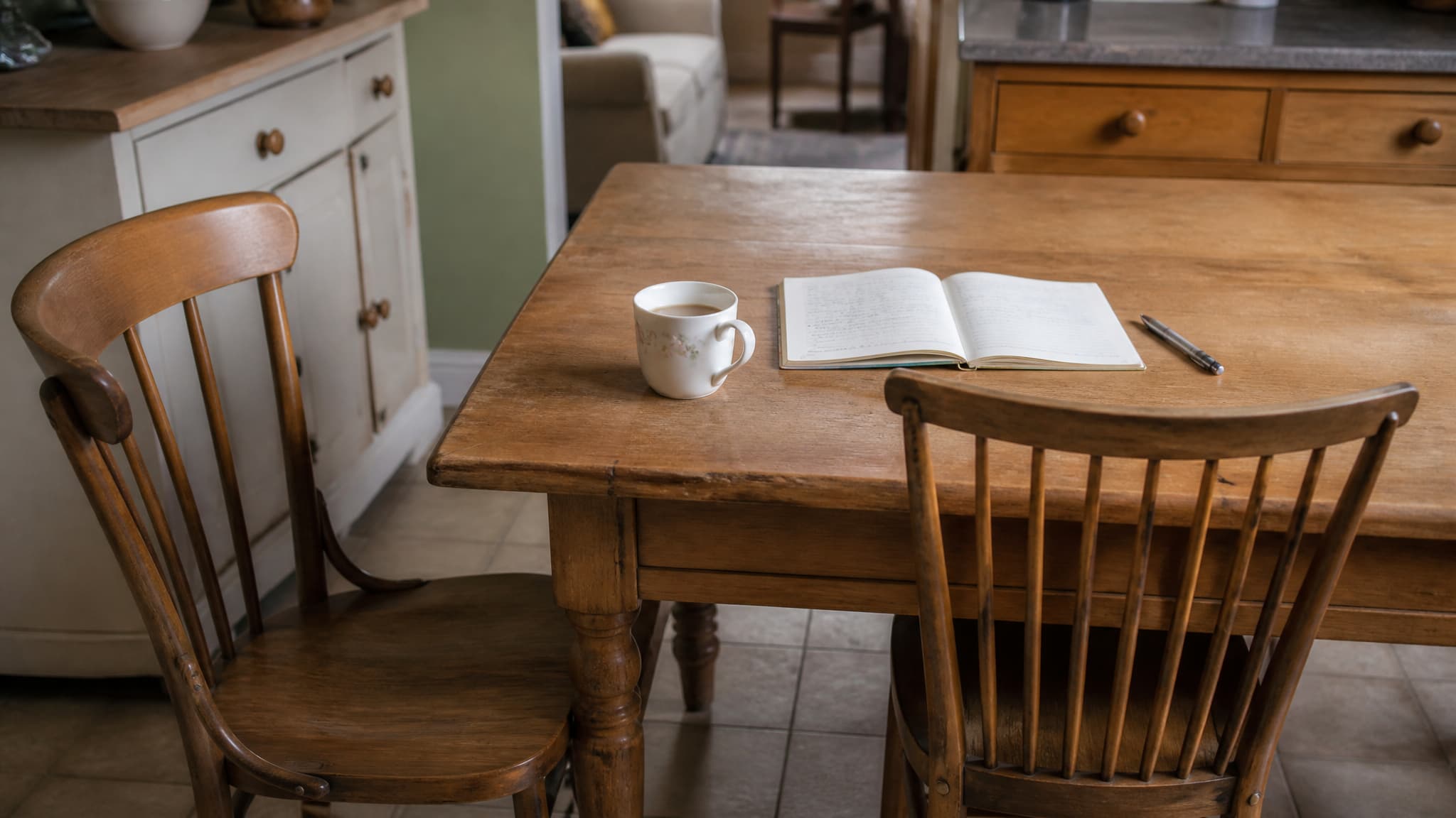 A kitchen table with two wooden chairs, one pulled out and turned away and one pushed in, a mug of tea and an open handwritten notebook beside a pen in flat afternoon light
