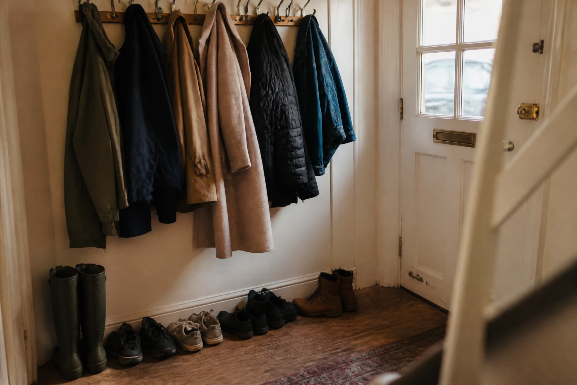 A row of coats on wall hooks by a front door, one of them a woman's wool coat still hanging among the others, boots below in a loose line