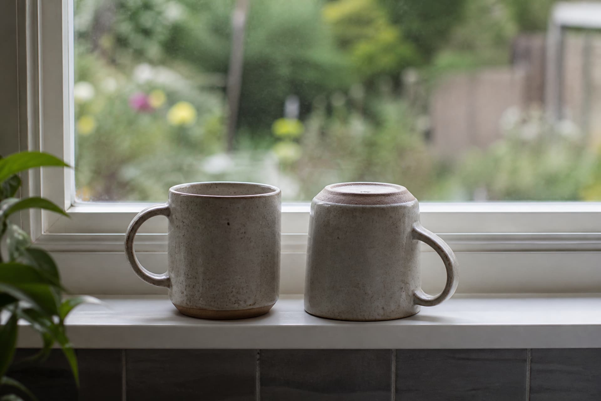 Two mugs on a kitchen windowsill in flat morning light, one upright and one turned upside down beside it, a garden out of focus through the glass