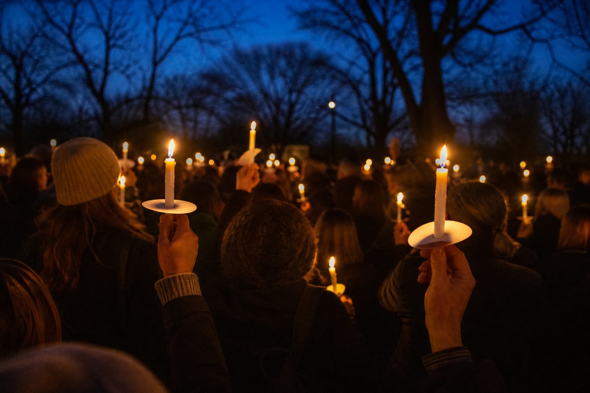 A crowd holding lit candles in paper drip protectors at dusk in a park, faces indistinct in the low warm light