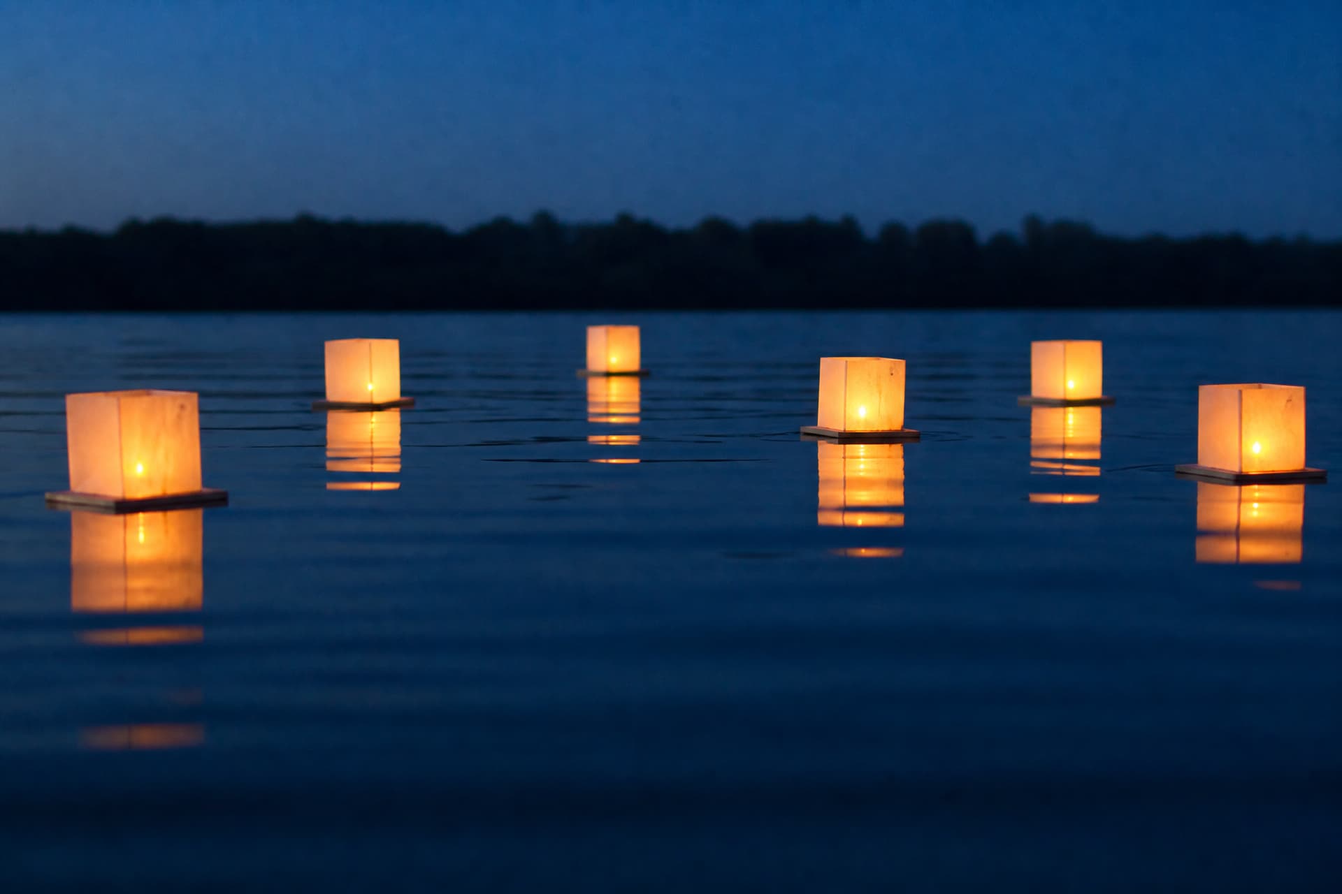 Small floating paper lanterns with candles drifting on still dark water at dusk, their reflections doubling the light