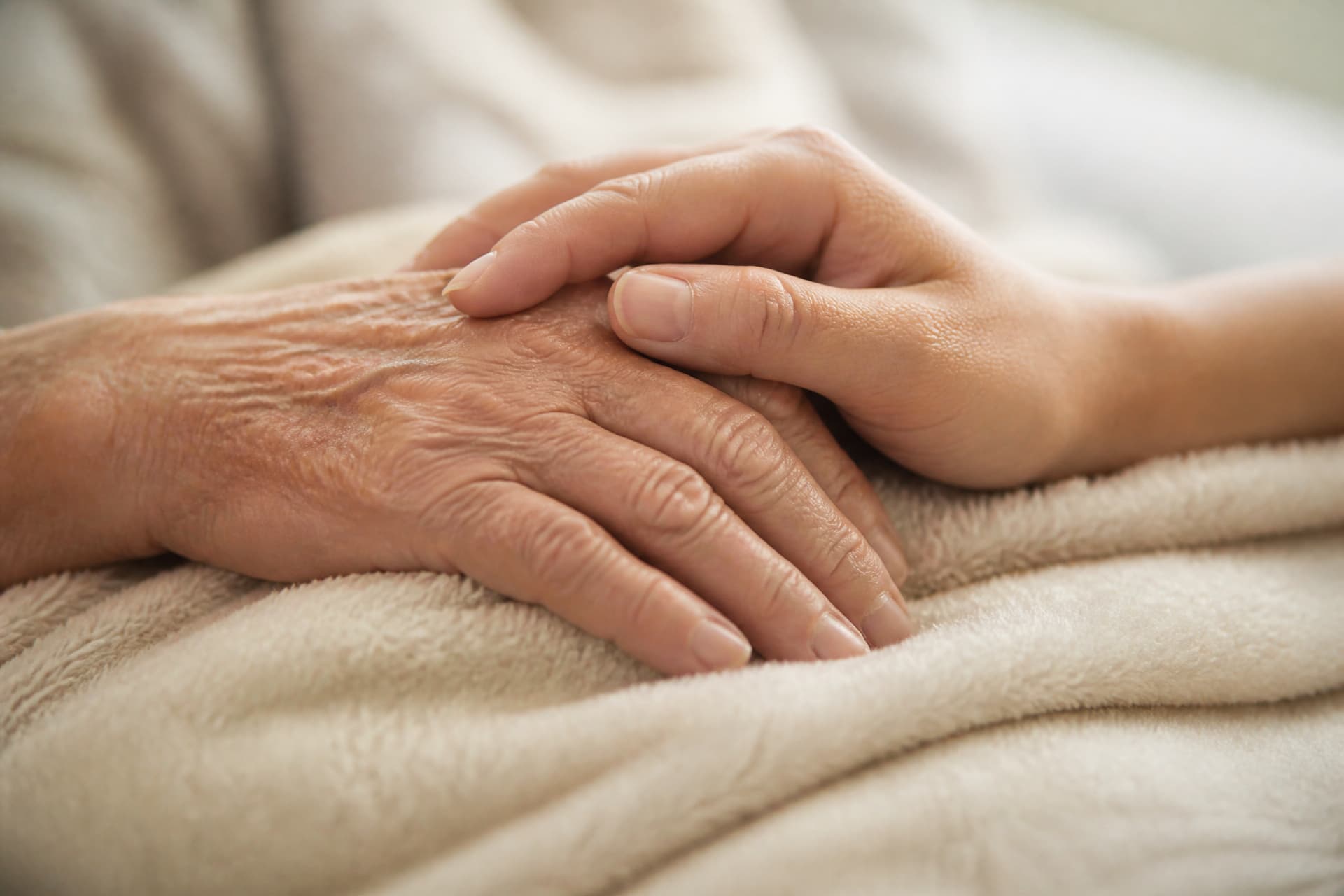 A younger hand resting gently over an older hand on the edge of a soft cream blanket