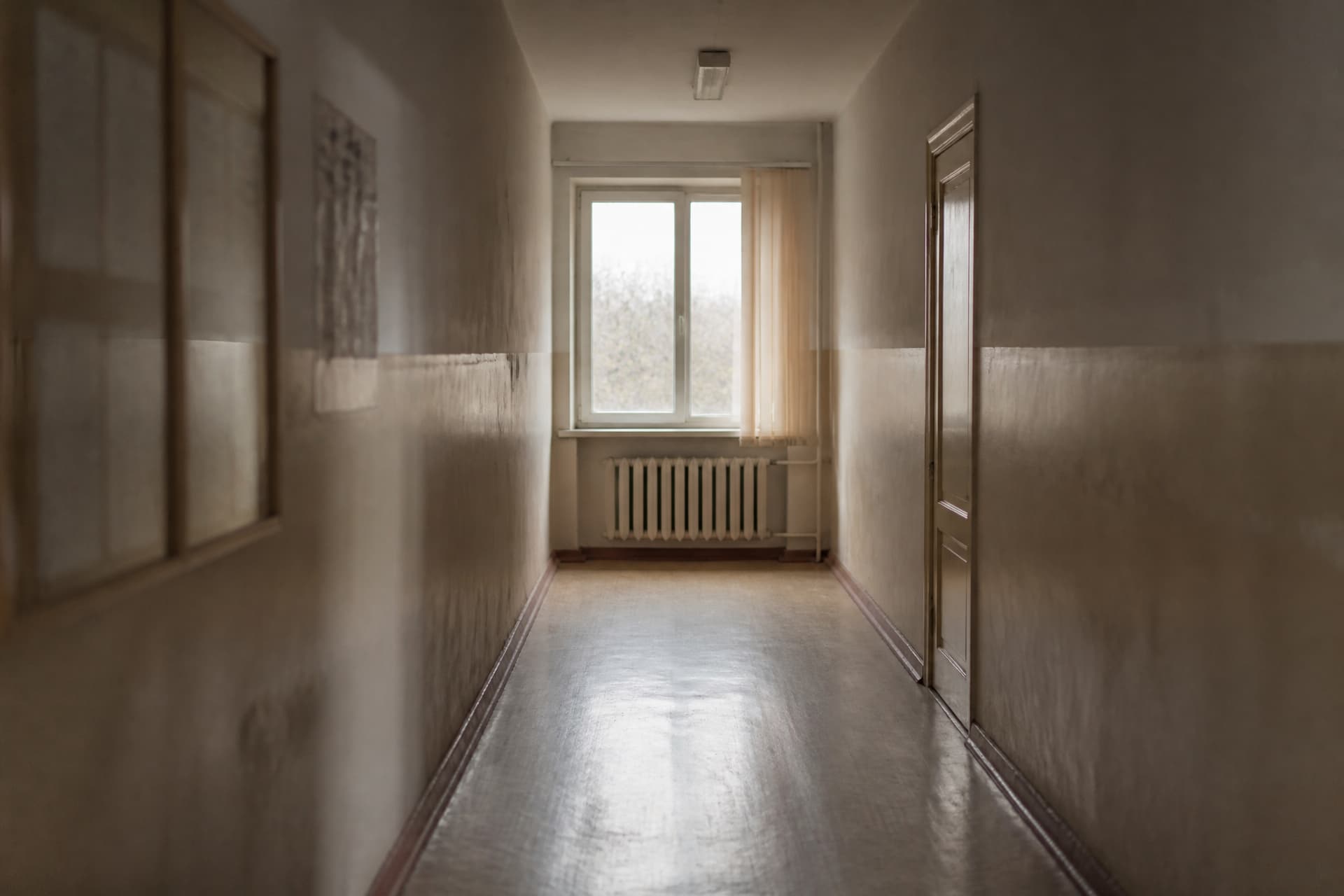 A quiet institutional corridor with a window at the far end and a single closed door, in flat overcast light