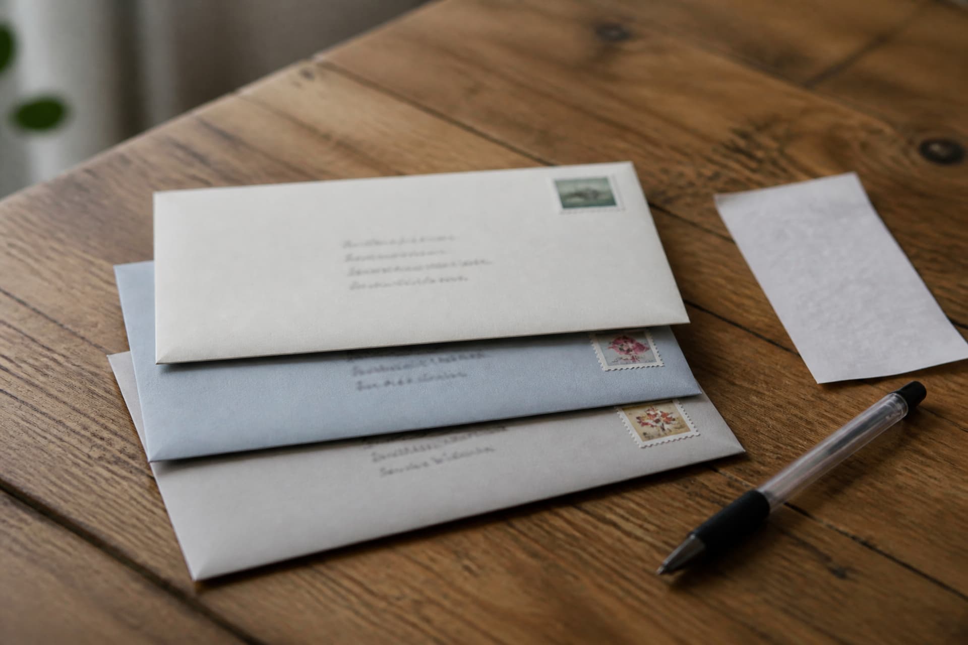 Three addressed envelopes stacked on a wooden table beside a certified mail receipt slip and a ballpoint pen