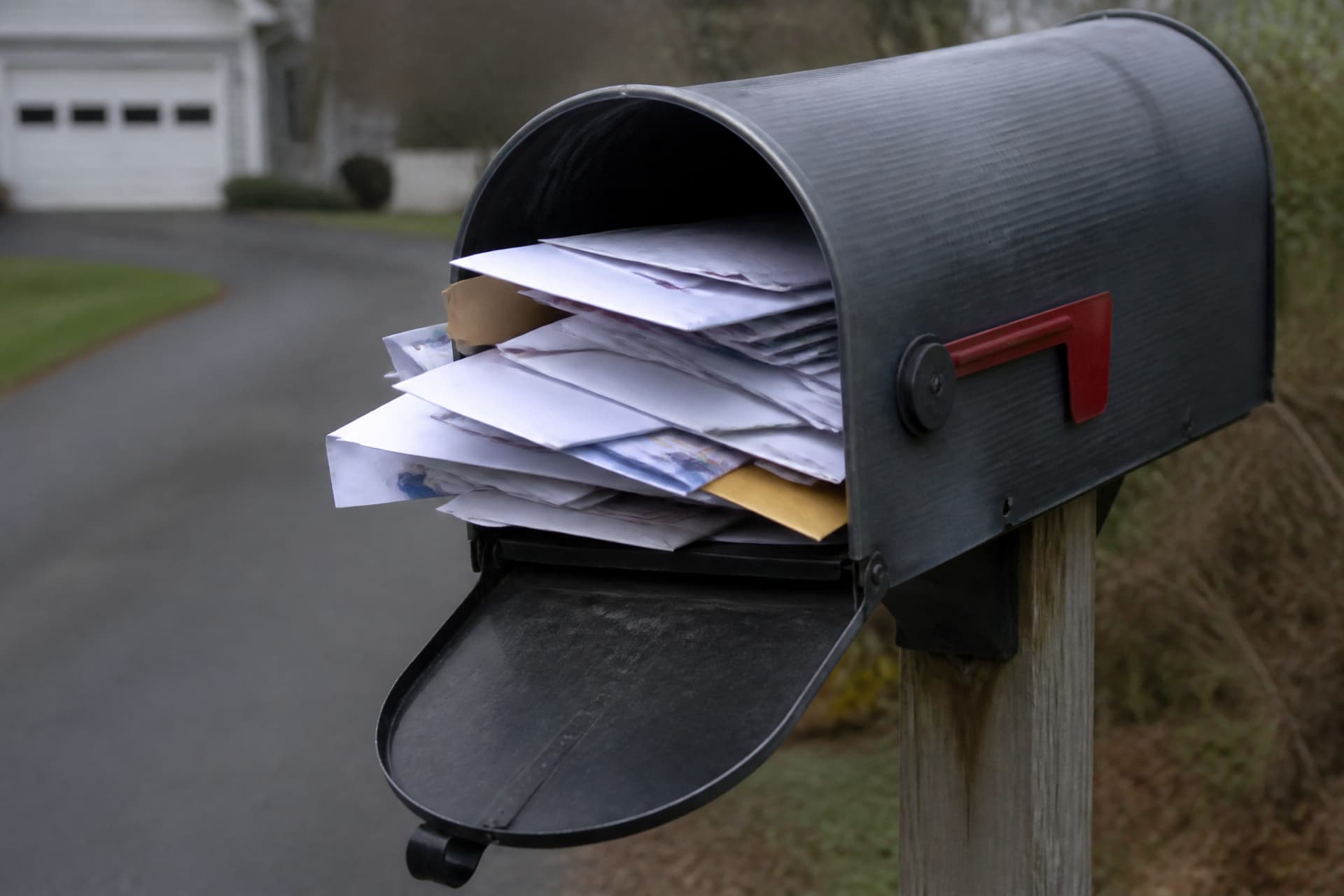 An overfilled residential mailbox at the end of a driveway with envelopes protruding, soft overcast light