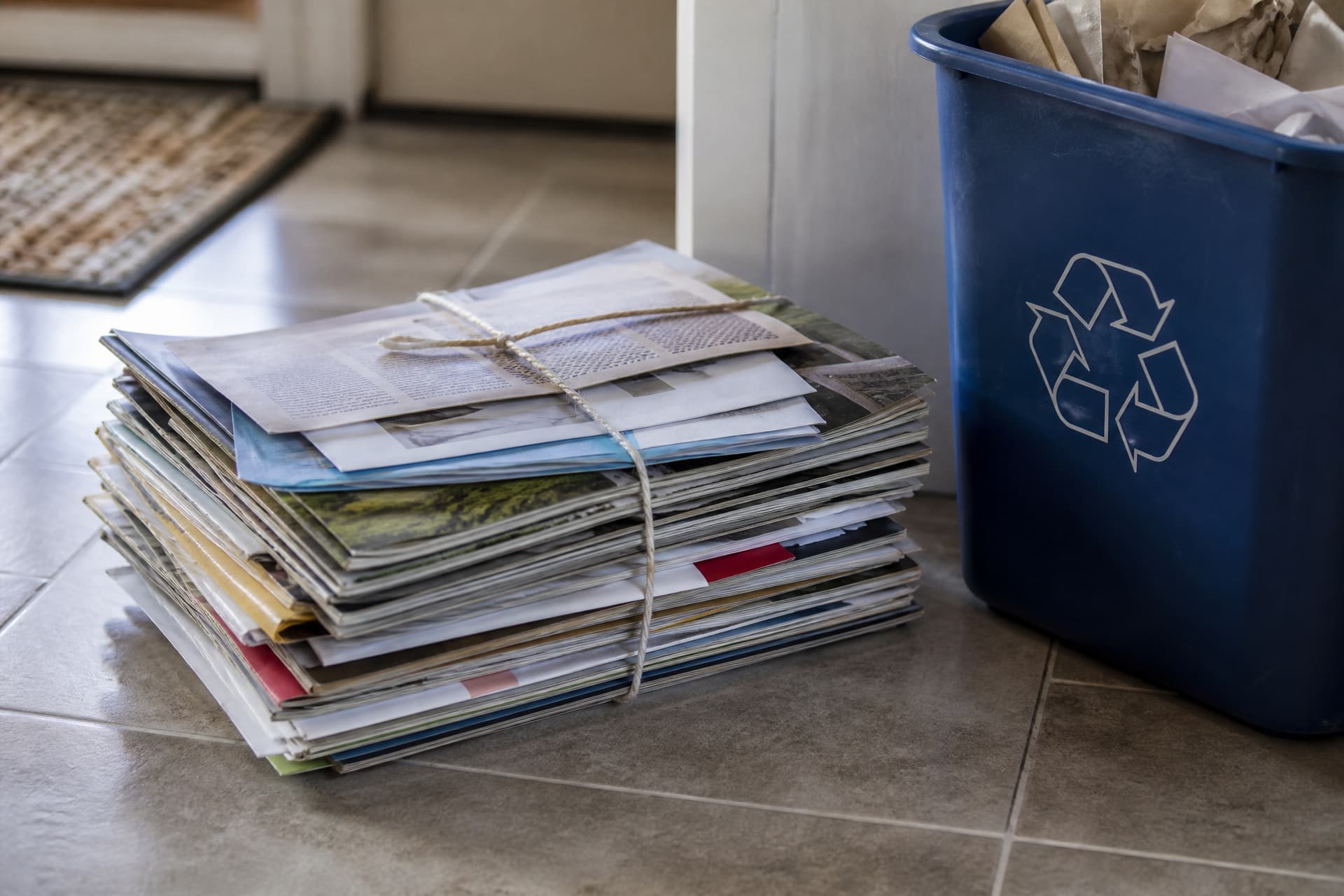 A stack of mail-order catalogs and promotional envelopes tied with string, set beside a recycling bin