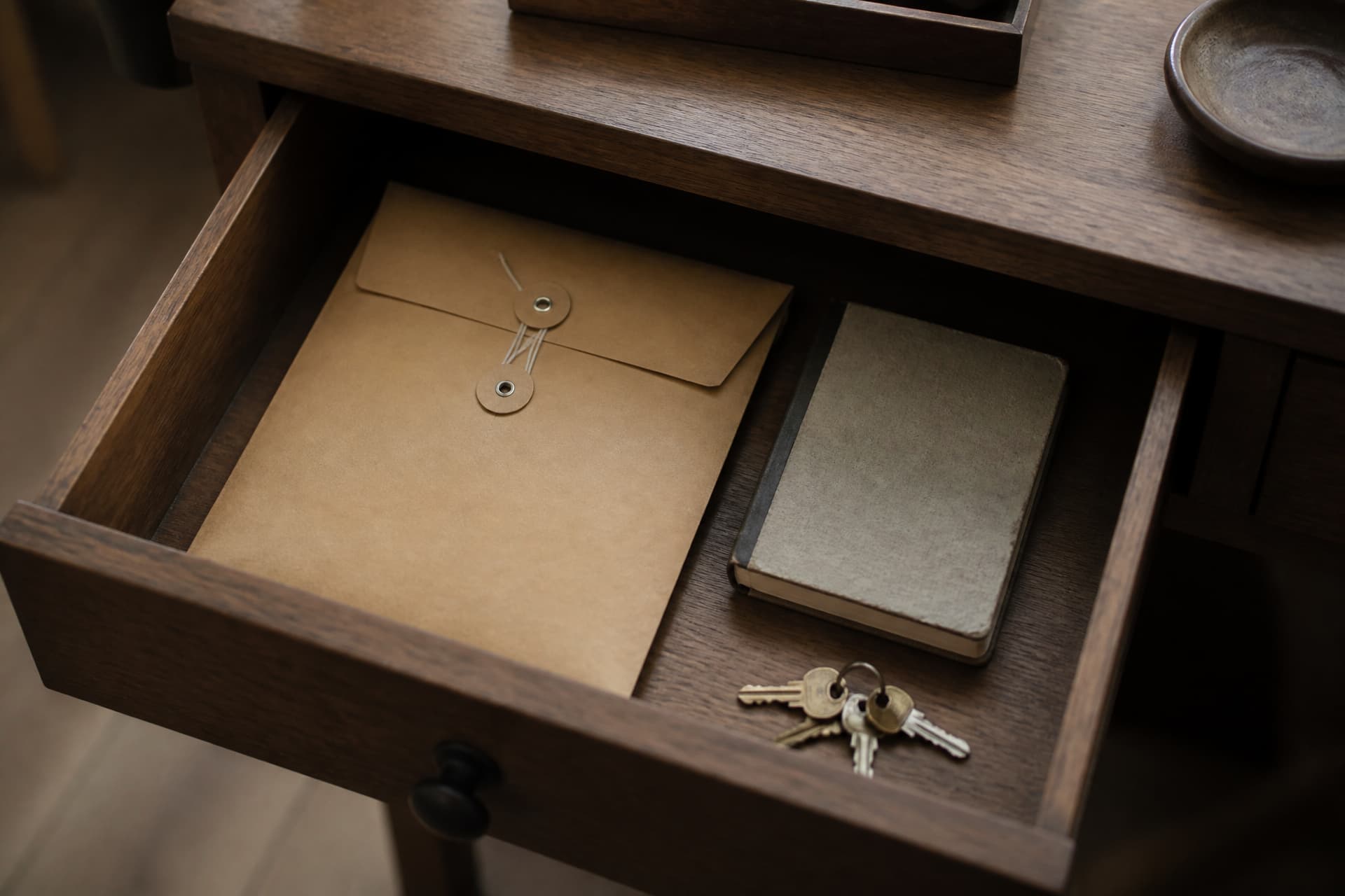 An open desk drawer containing a large sealed envelope, a small notebook and a set of spare keys, seen from above in warm light