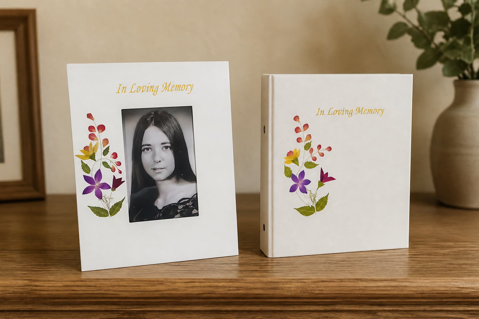 A cream memorial picture frame holding a black and white portrait and a matching memorial photo album, both decorated with pressed flowers, standing on a wooden shelf
