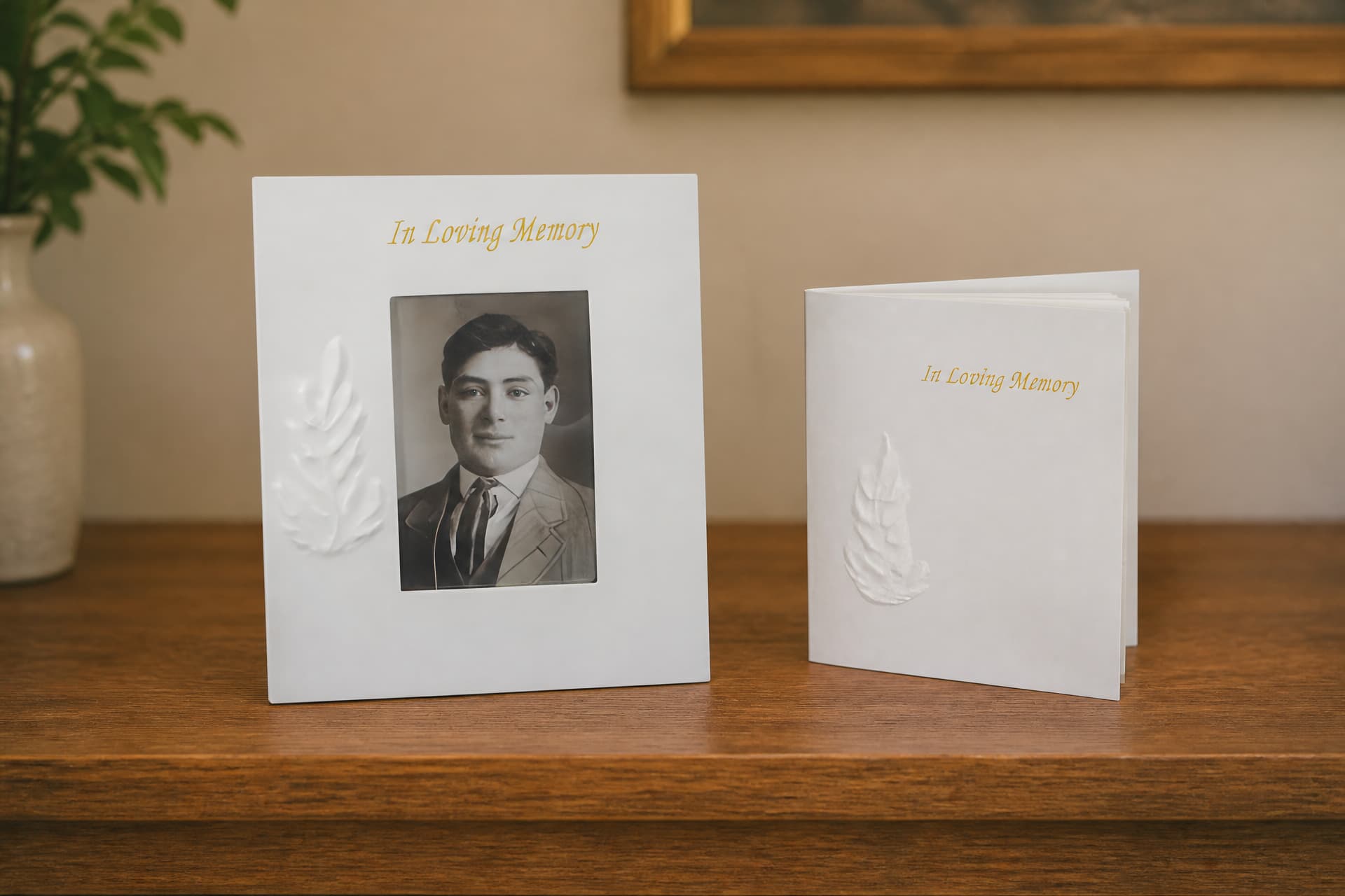A white memorial picture frame holding a vintage portrait beside a matching memorial guest book, both embossed with a leaf motif, standing on a wooden shelf
