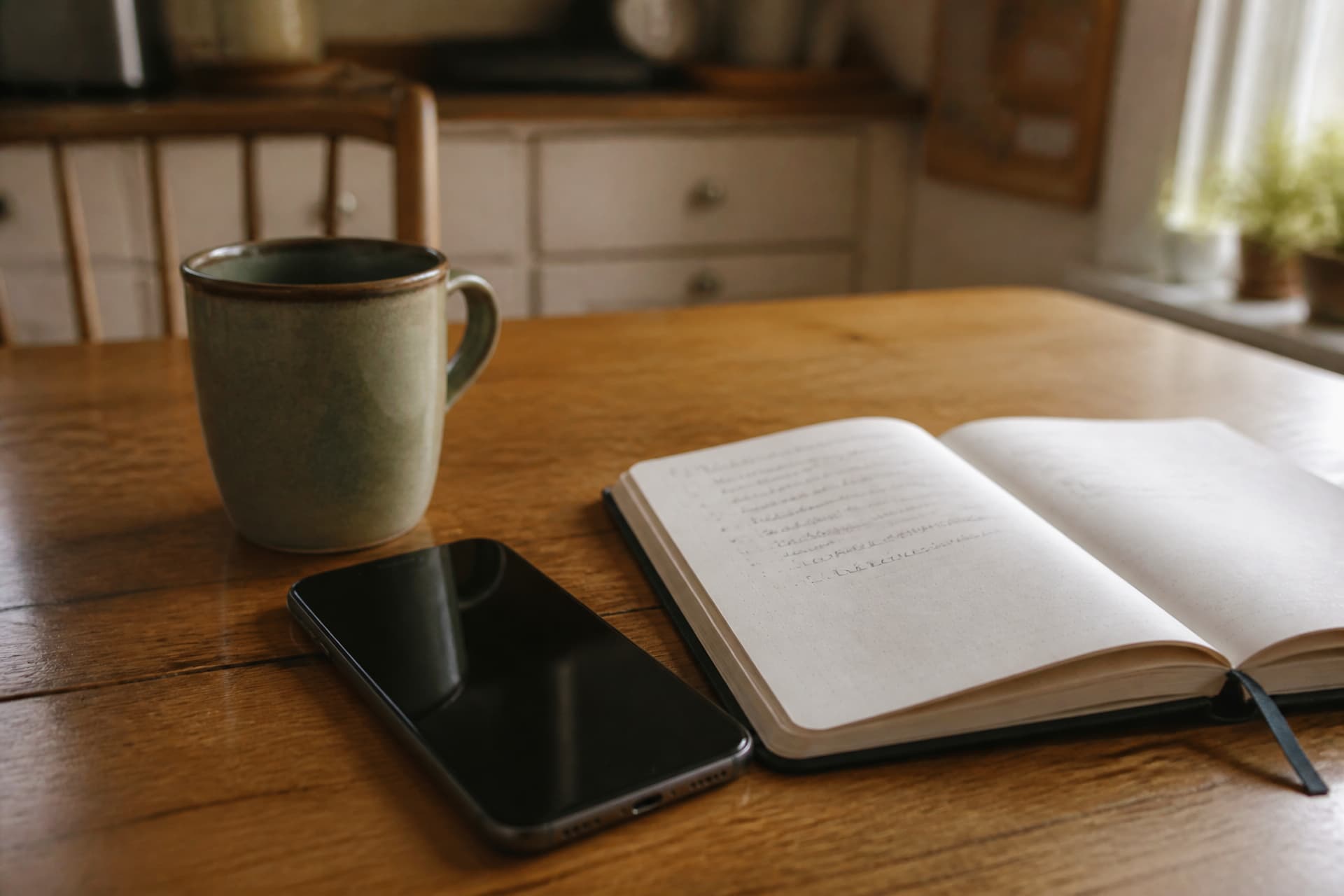 A phone face-up on a kitchen table beside an open notebook with a short handwritten list and a mug, warm daylight