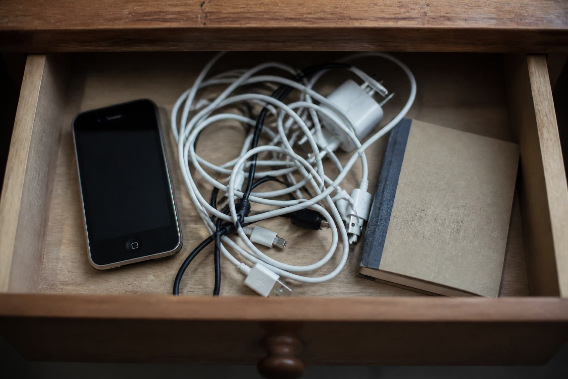 An open drawer holding an older phone, a tangle of charging cables and a small notebook, seen from above in flat light