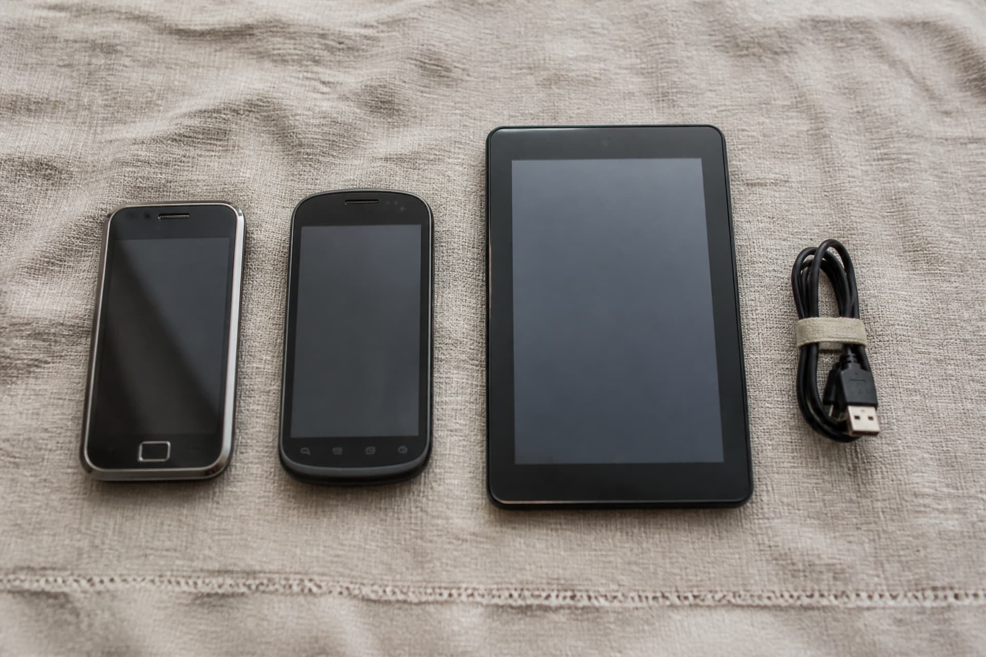 Two older smartphones and a small tablet arranged on a linen cloth beside a folded charging cable, flat overhead light