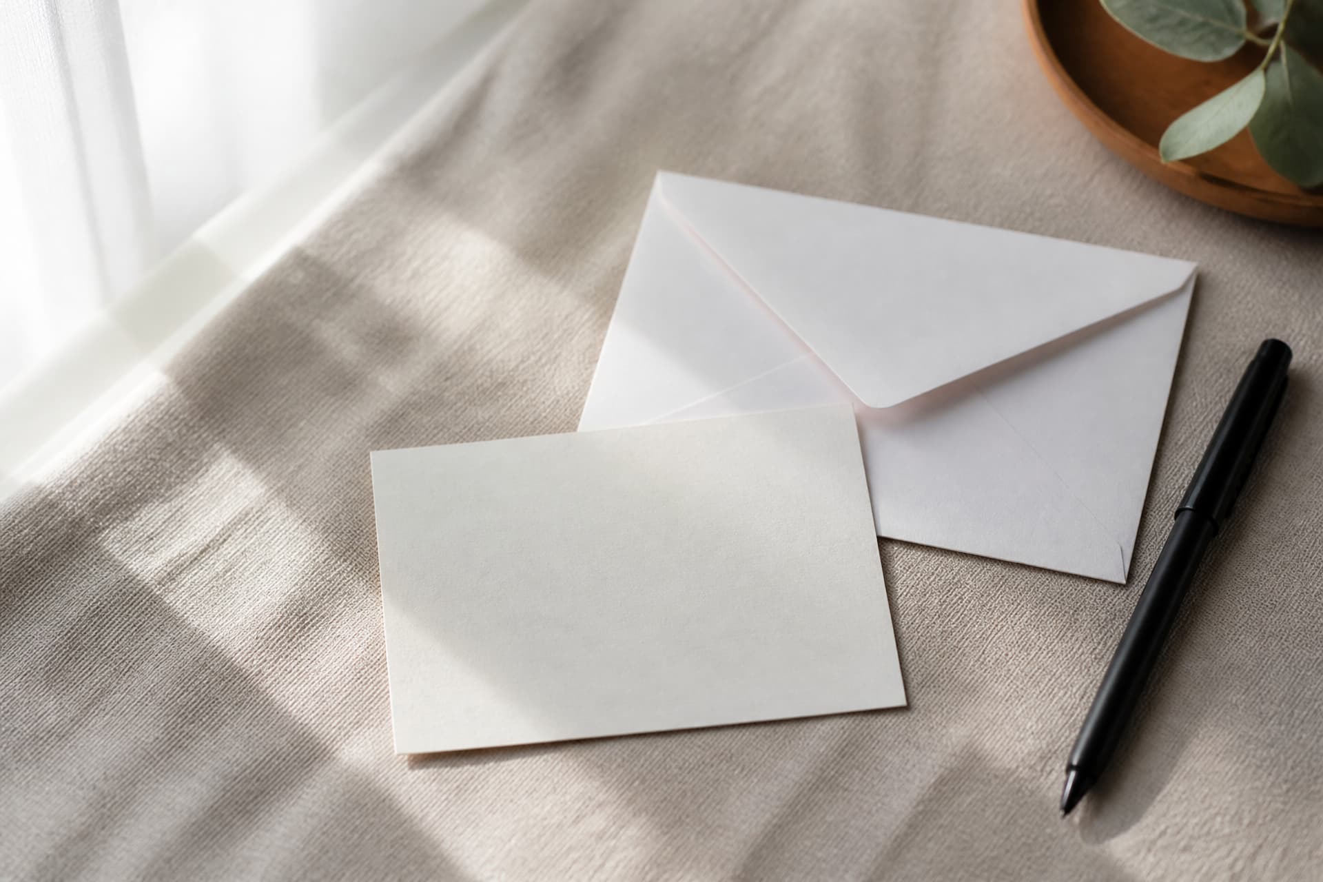 A small blank note card and a pen resting on a linen tablecloth beside a white envelope, photographed from above in soft daylight