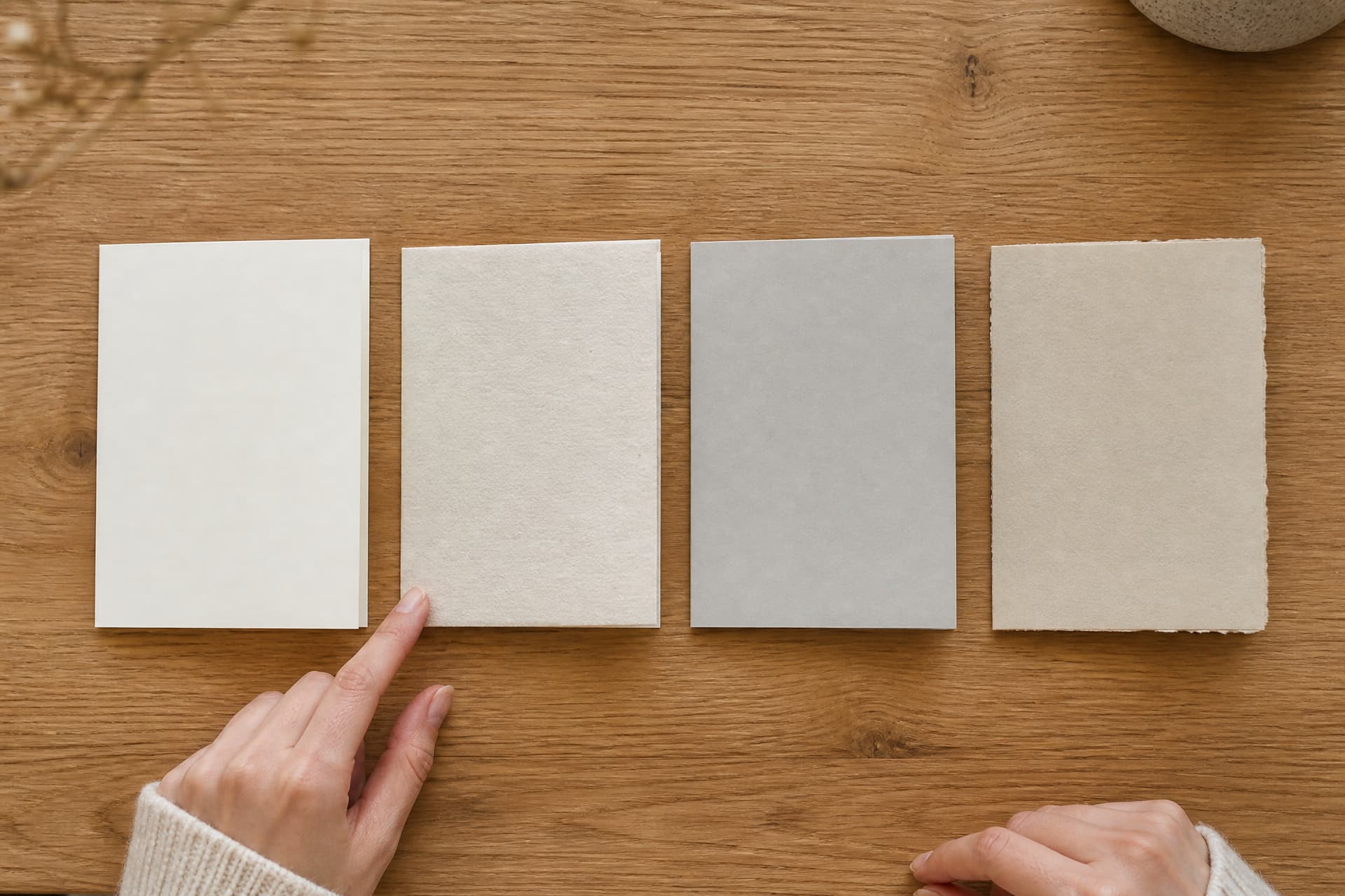 Four blank folded sympathy cards in different papers laid side by side on an oak table, a hand touching one