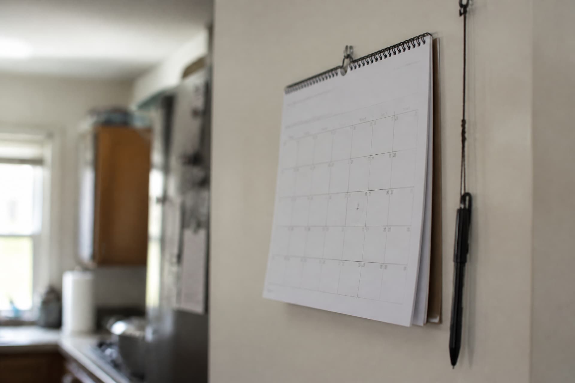 A paper wall calendar hanging in a kitchen showing a month as a grid of small squares, a single date ringed faintly in pencil, a pen on a string beside it