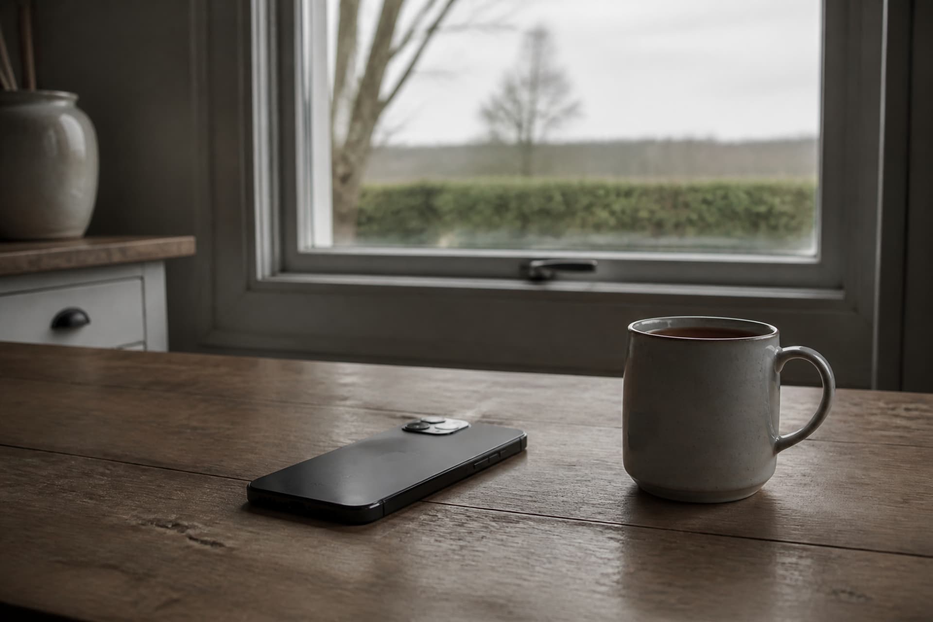 A phone lying face down on a wooden kitchen table beside an untouched mug of tea, flat grey daylight from a window behind